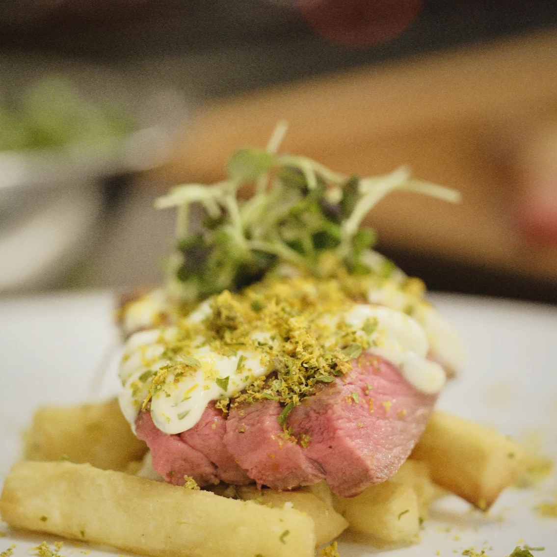 Close-up of gourmet dish with a piece of pink meat, topped with white sauce, crushed herbs, and microgreens, served on a bed of fries.