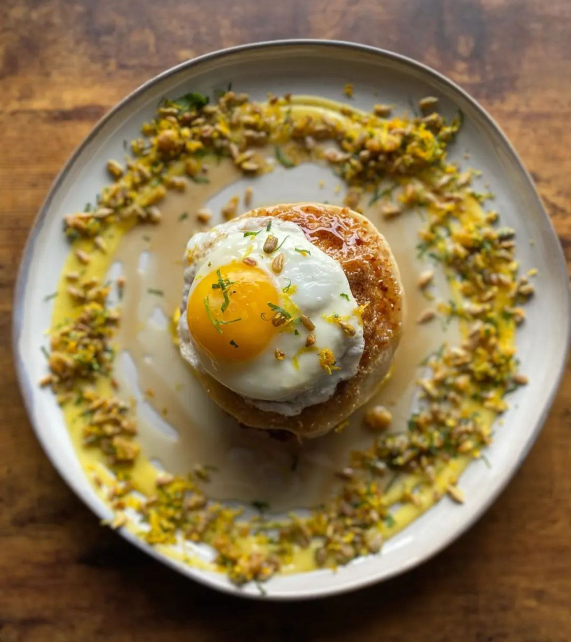 A plate with a fried egg on top of a biscuit or similar bread, garnished with lemon zest, surrounded by chopped pistachios and a drizzle of yellow sauce.