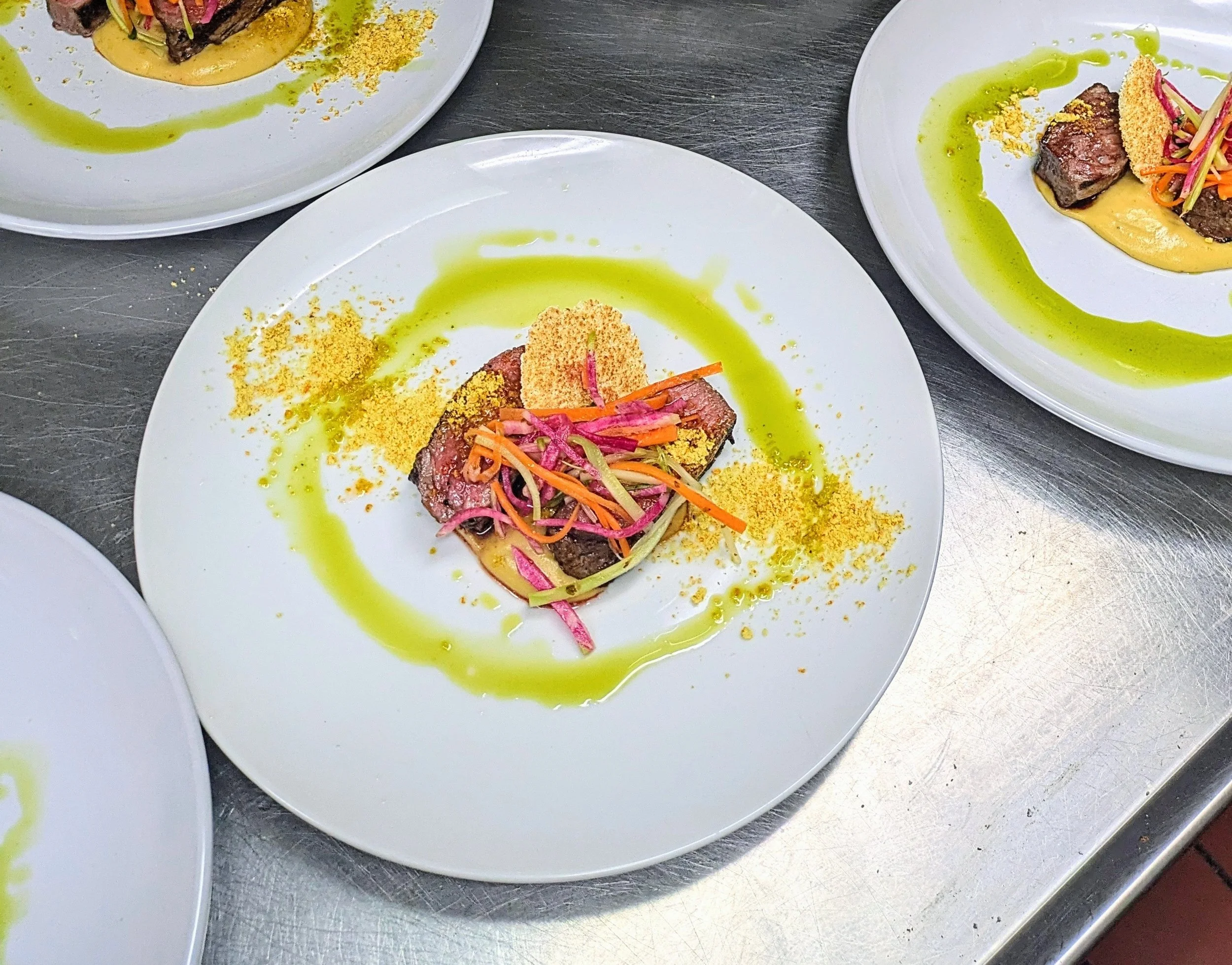 A plated dish featuring a small piece of medium-rare steak topped with colorful pickled vegetables, a crunchy chip, and a drizzle of green sauce, garnished with yellow crumbs and a smear of yellow puree.