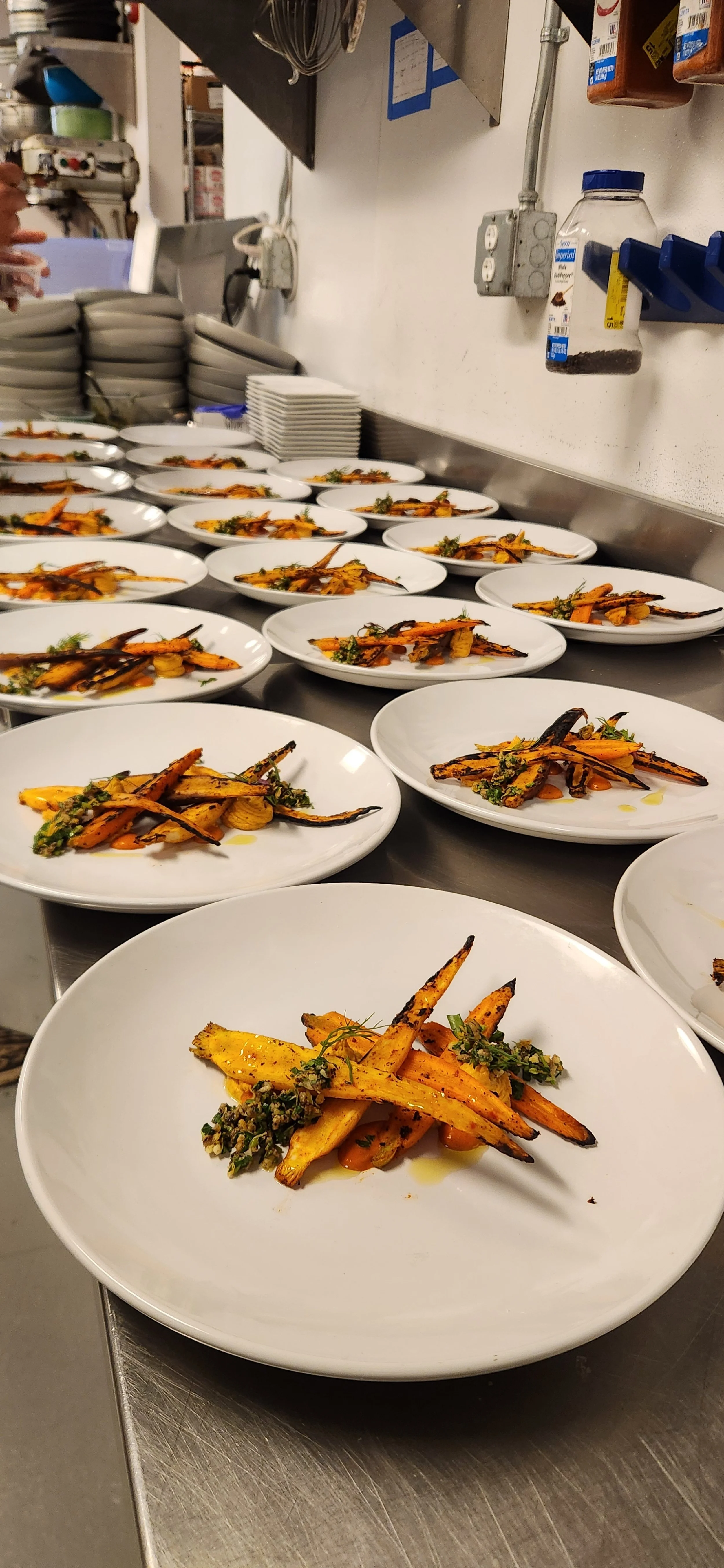Multiple white plates with roasted carrots and garnishes arranged on a stainless steel kitchen counter, with kitchen tools and dishes stacked in the background.