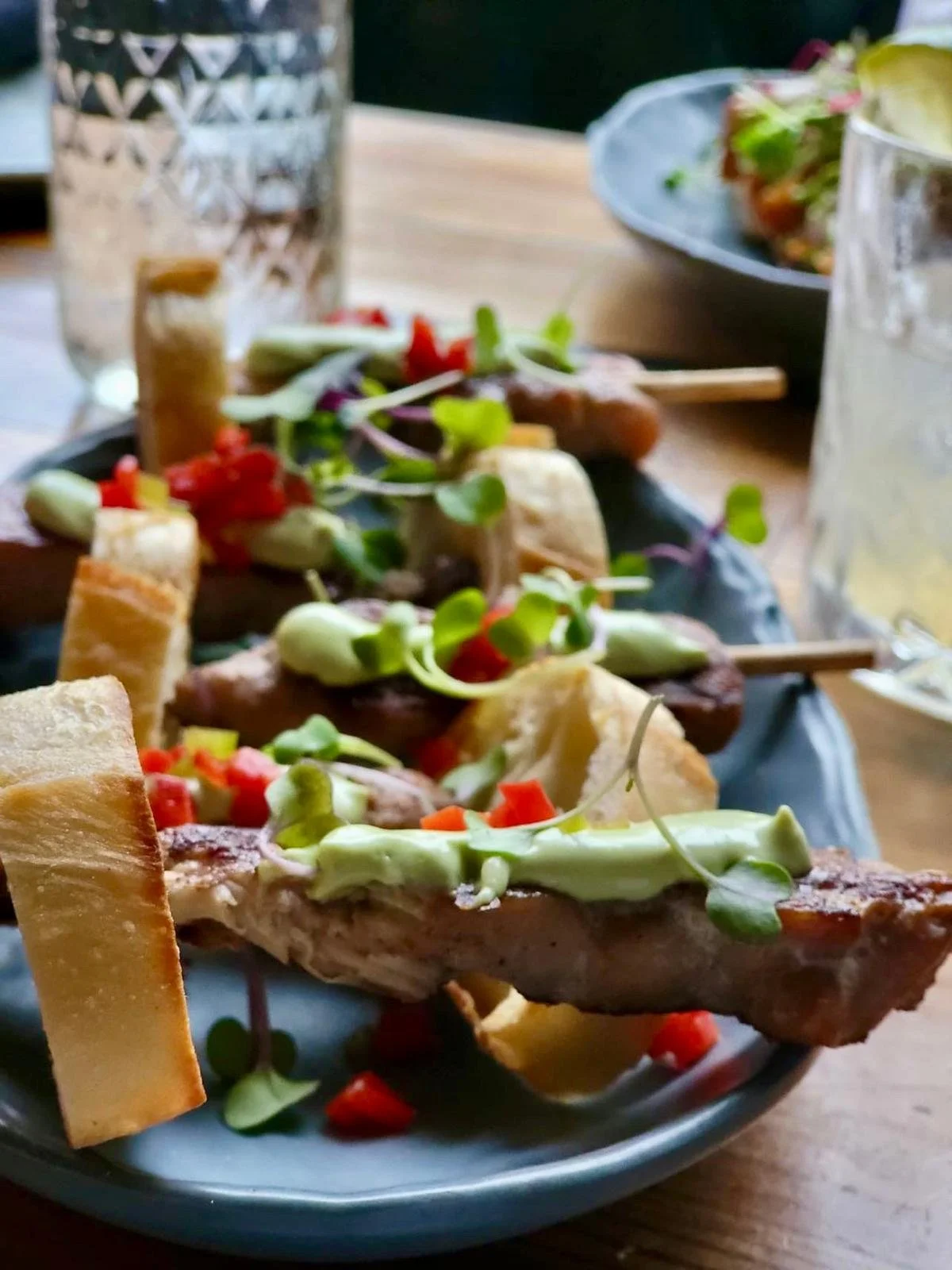 Close-up of a plate of appetizers with bread, green sauce, diced red peppers, microgreens, on skewers, on a dark ceramic plate on a wooden table, with a glass of water and another dish in the background.