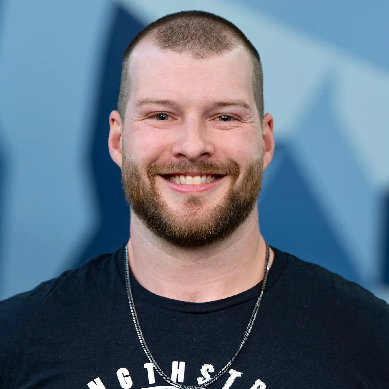 A smiling man with a beard and short hair, wearing a black T-shirt and a necklace, standing in front of a blue background.