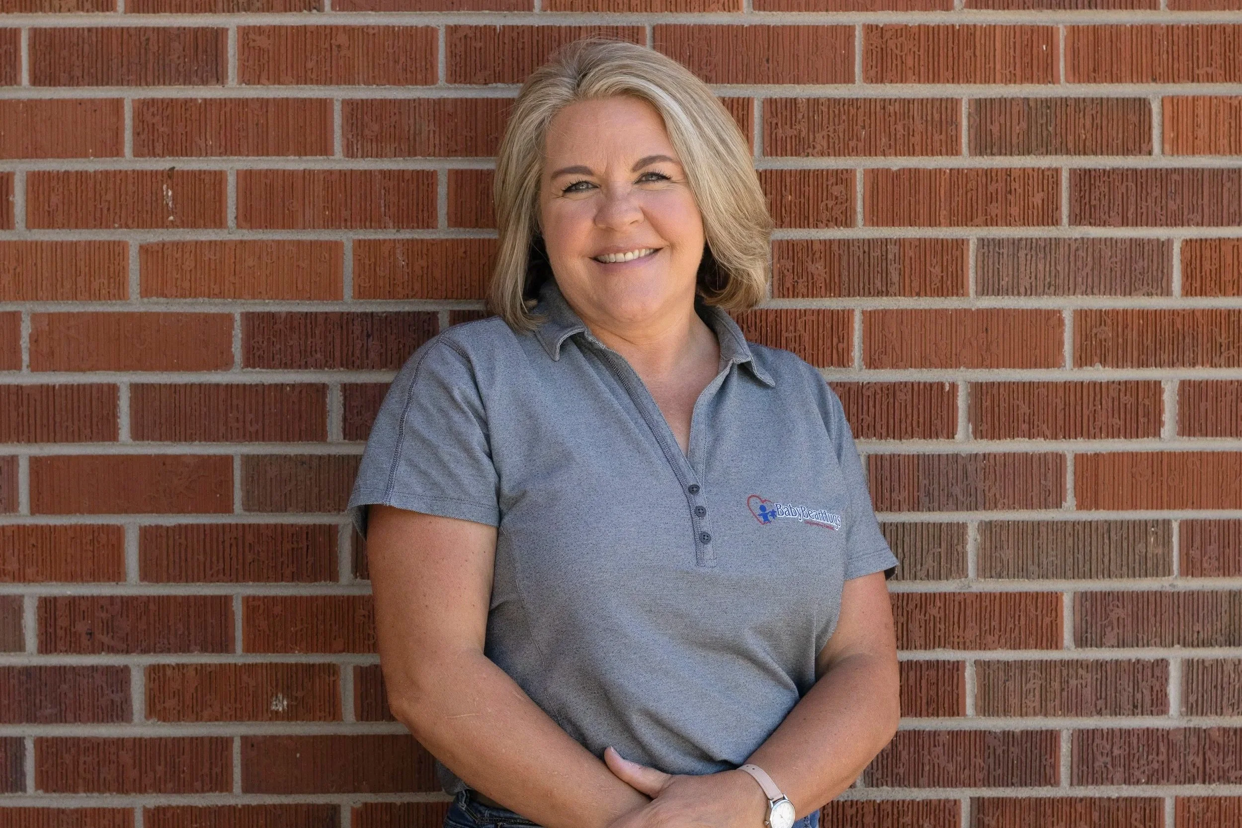 Smiling woman with blonde hair standing against a brick wall, wearing a gray polo shirt with a logo.
