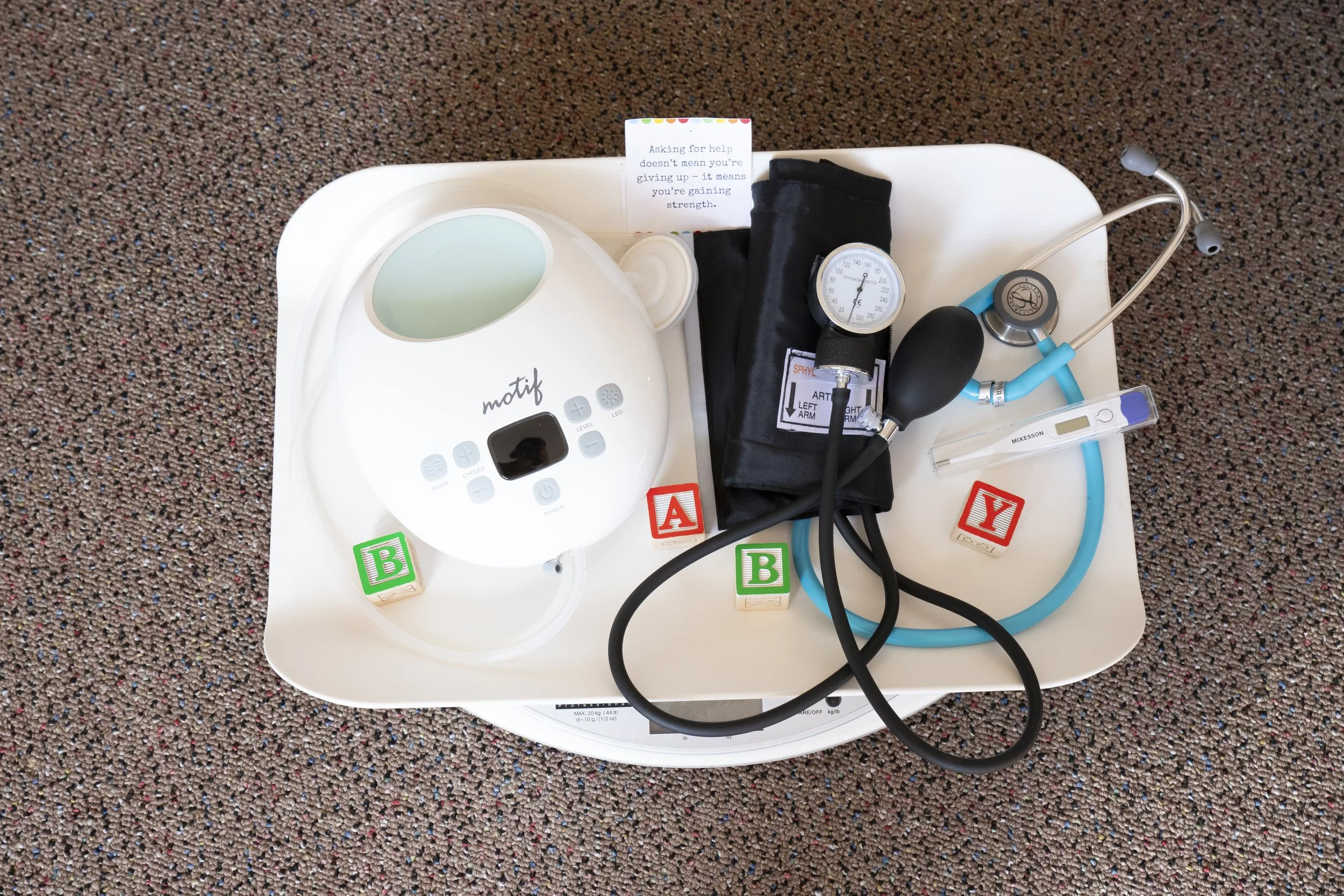 Medical supplies on a tray, including a blood pressure cuff, a thermometer, a stethoscope, and a digital monitor, with color-coded letter blocks reading B, A, Y, and B.