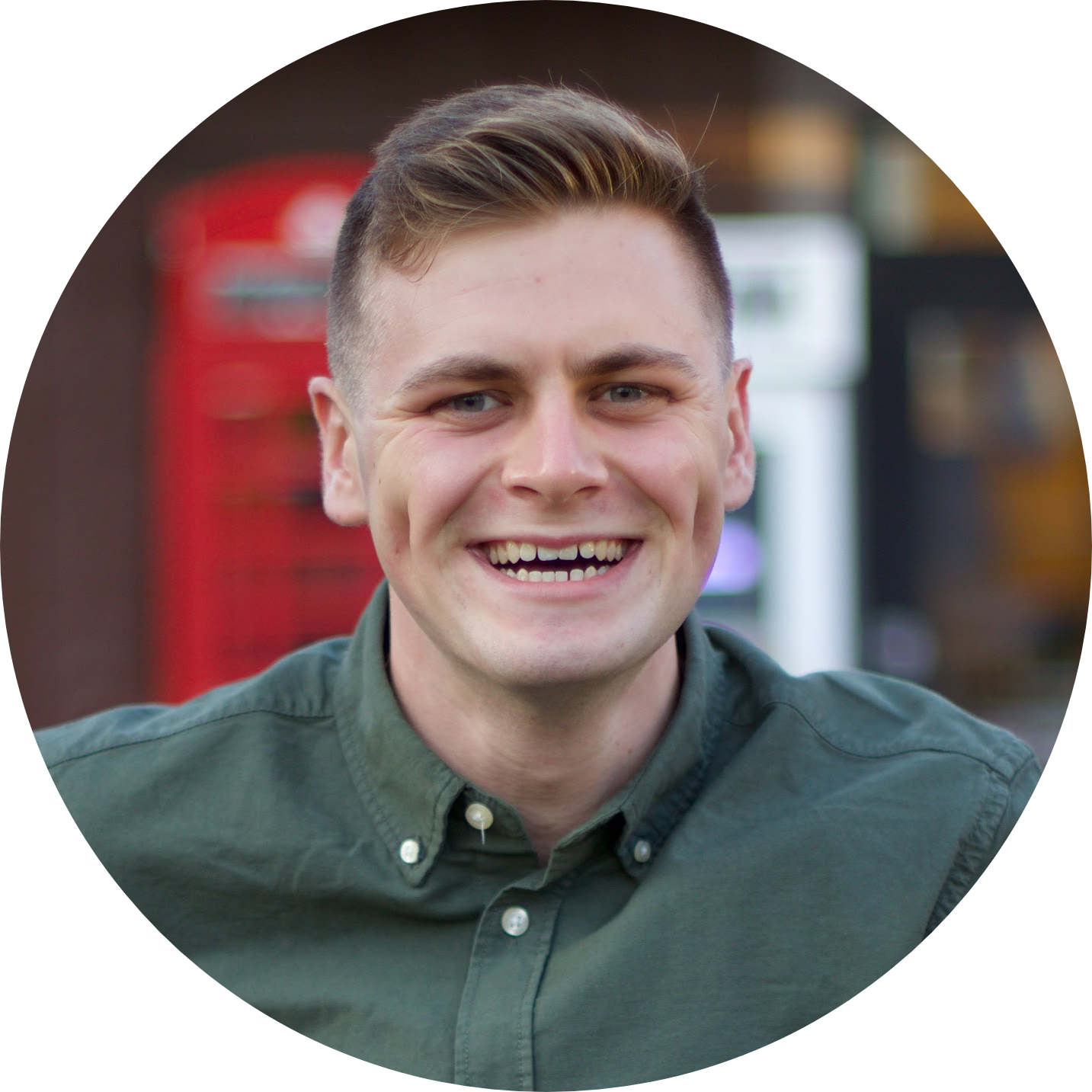 A young man with short, light brown hair smiling, wearing a dark green button-up shirt. Background includes blurred out red and black objects.