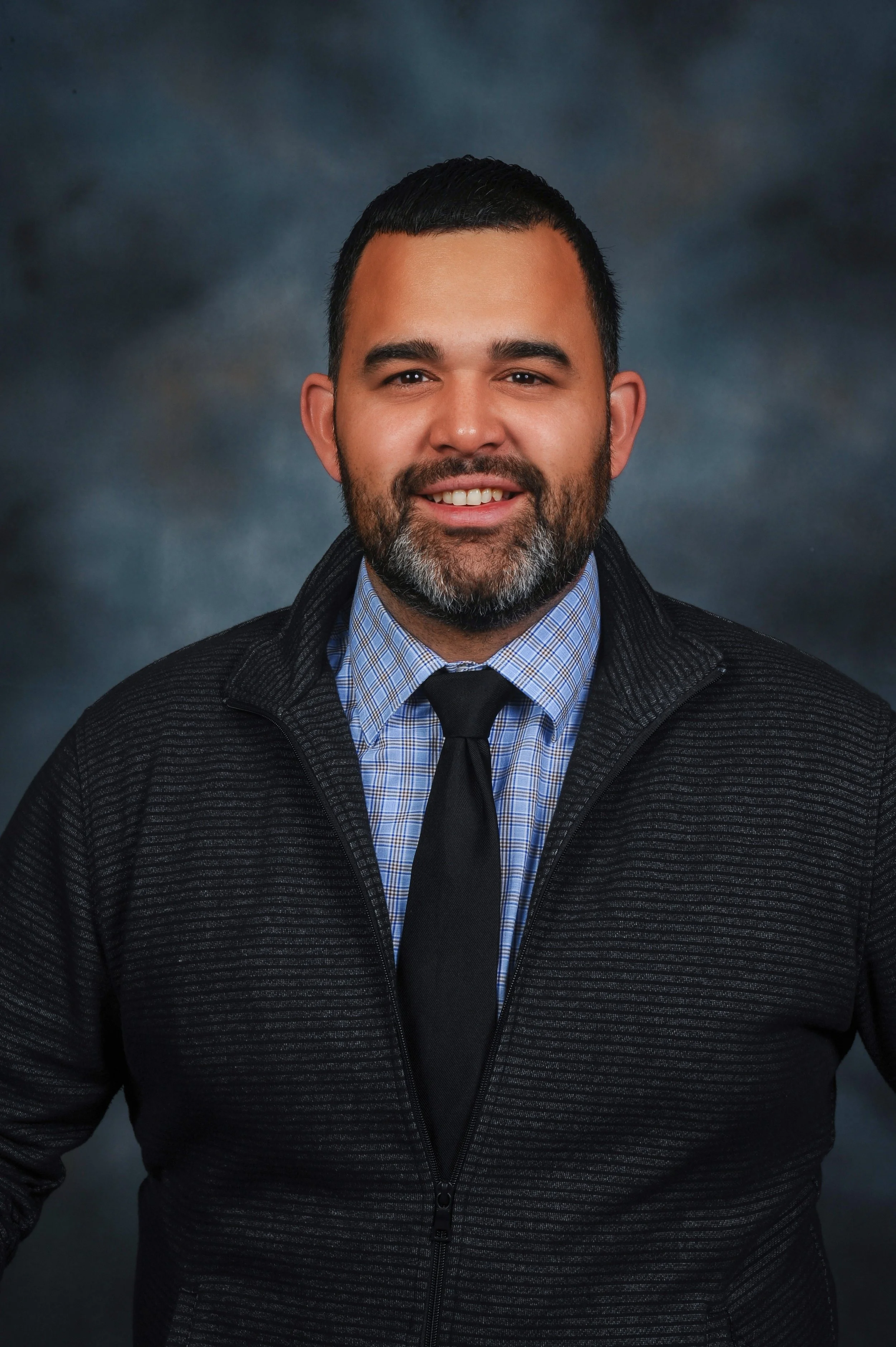 Headshot of a man with dark hair and beard, wearing a dark textured jacket, a blue checkered shirt, and a black tie, smiling against a dark cloudy background.