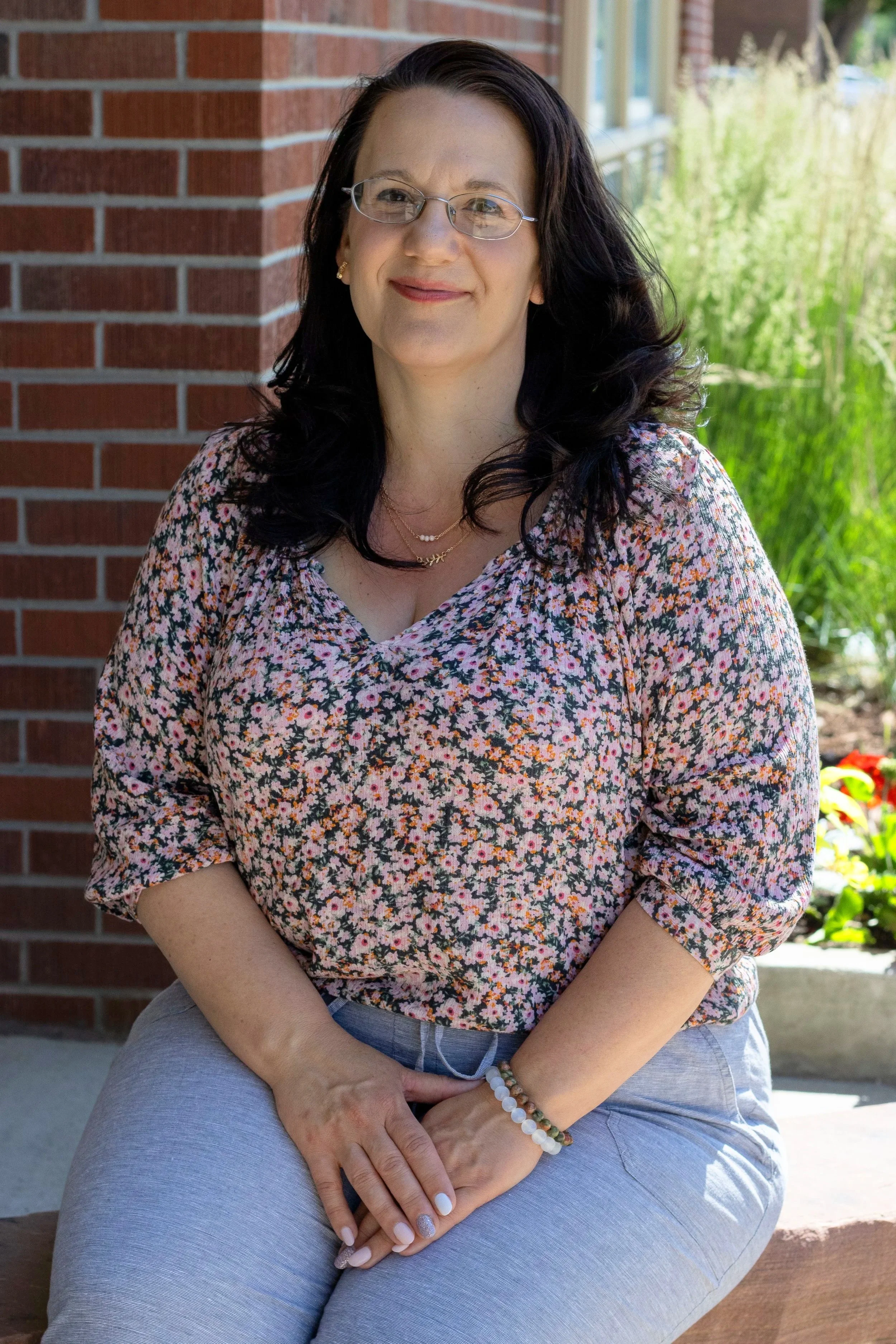 A woman with dark wavy hair, glasses, and light skin sitting outdoors near a brick wall next to a garden.