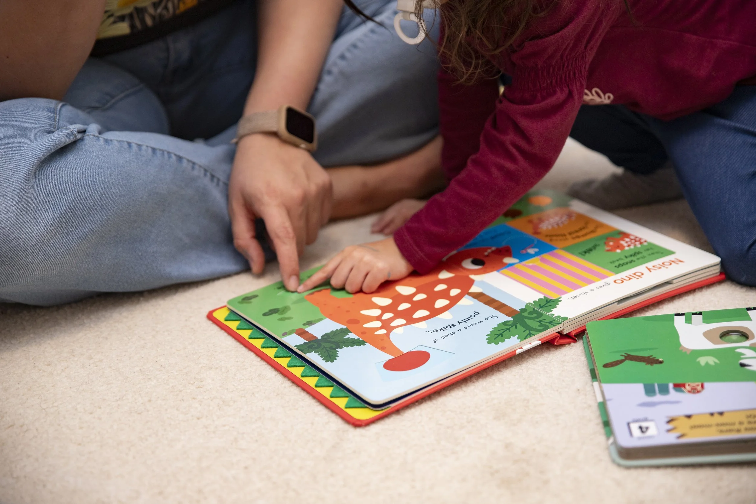 A child and an adult sitting on a carpet, looking at a colorful children's book about animals, with the child pointing to the illustrations.