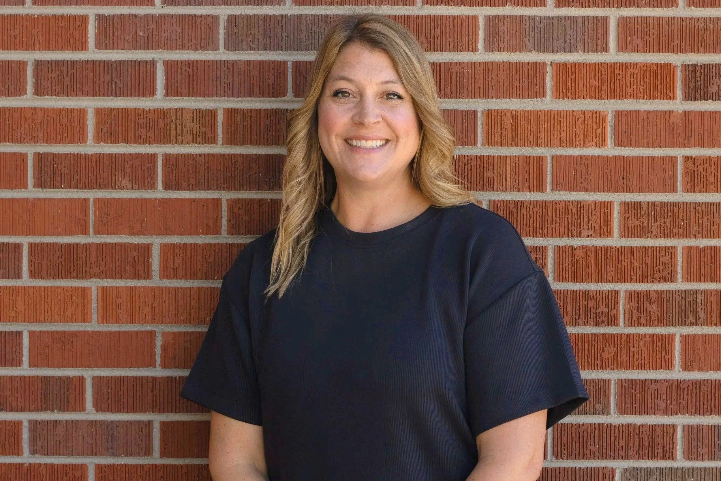 A smiling woman with blonde wavy hair wearing a black t-shirt, standing against a brick wall.