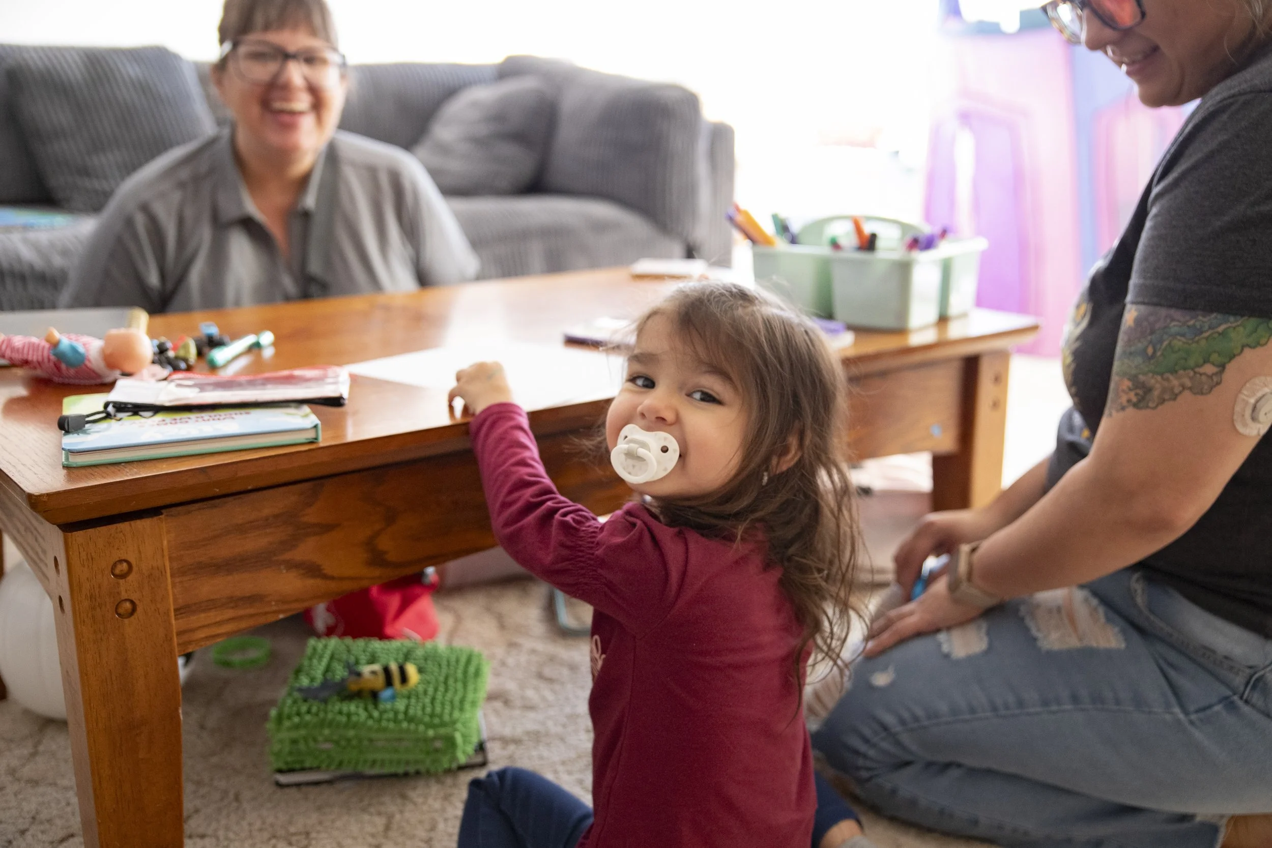 A young girl with a pacifier in her mouth, sitting on the floor, looking up at the camera. She is at a living room table with a woman and another adult nearby. The table has toys, books, and art supplies. The girl looks curious and is reaching for something on the table.
