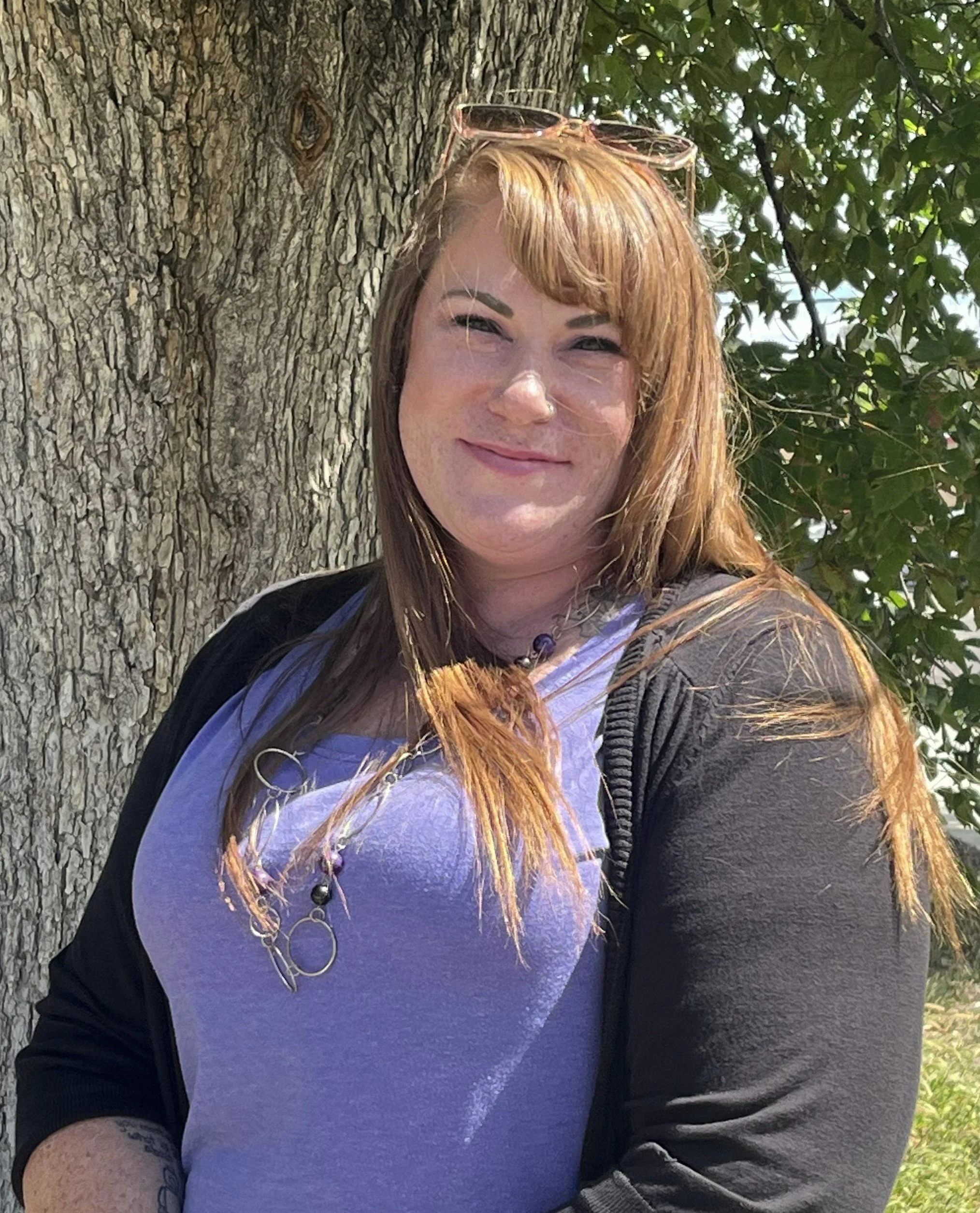 A woman with long brown hair, wearing a white blazer and black shirt, sitting outdoors on a stone surface with a grassy lawn and trees in the background.