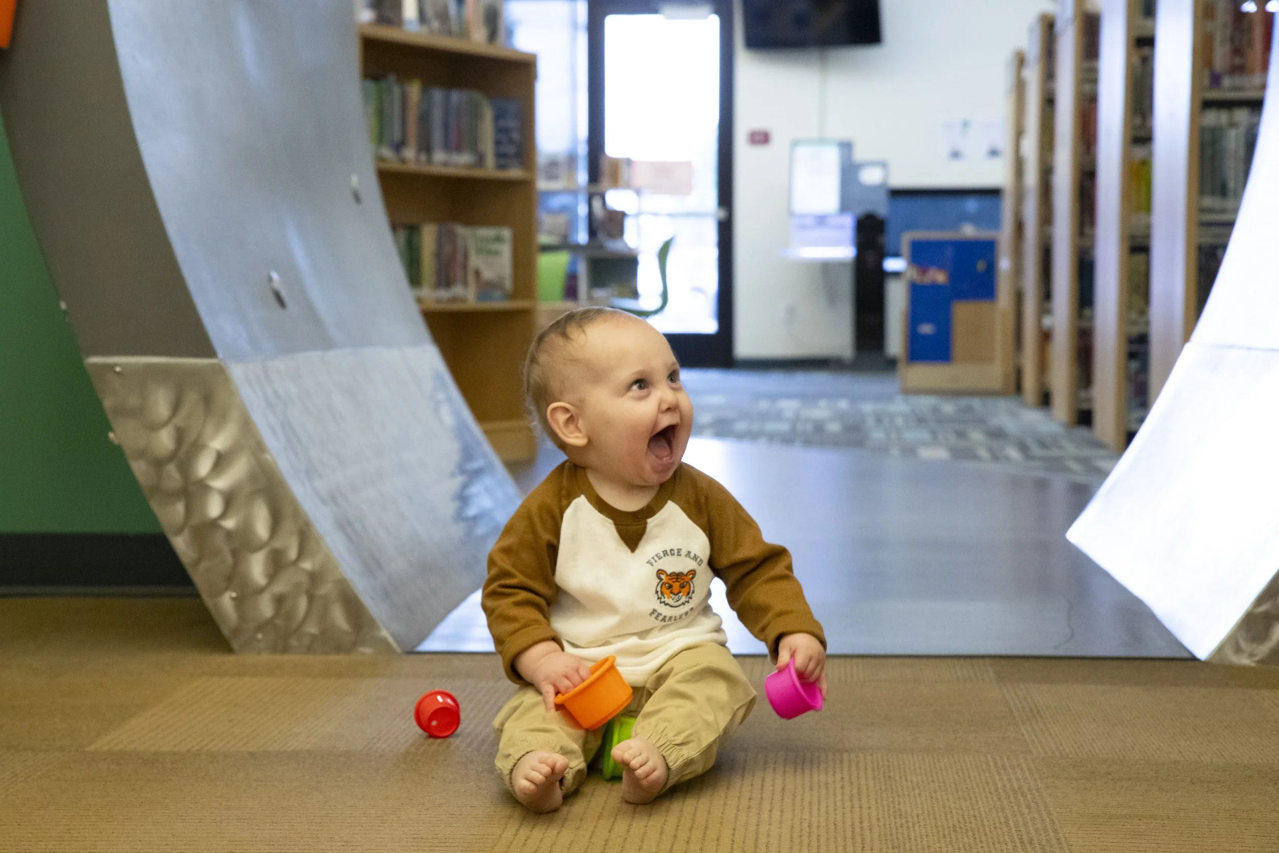 A baby sitting on the floor in a library or bookstore, smiling and holding colorful cups.
