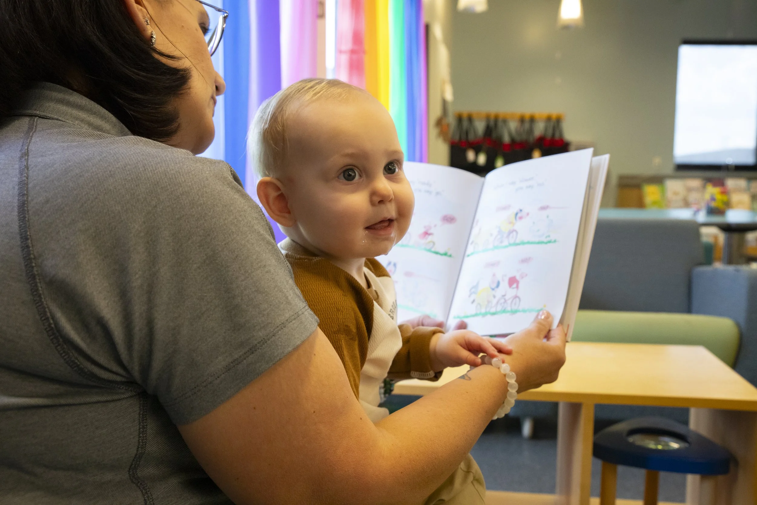 A woman holding a young boy reading a colorful picture book inside a room with rainbow curtains and colorful decorations.