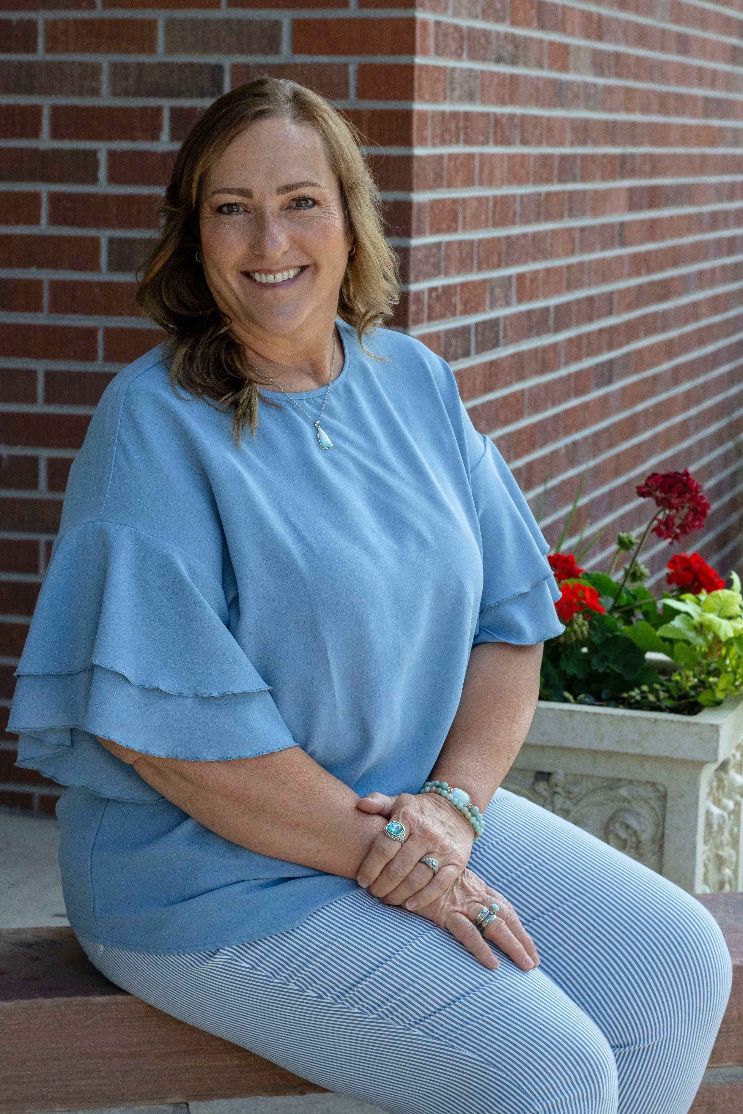 A woman with light brown hair sitting on a bench in front of a red brick wall. She is wearing a light blue blouse with ruffled sleeves, striped pants, and jewelry, and is smiling while looking at the camera. There is a white planter with red and green flowers on her right.