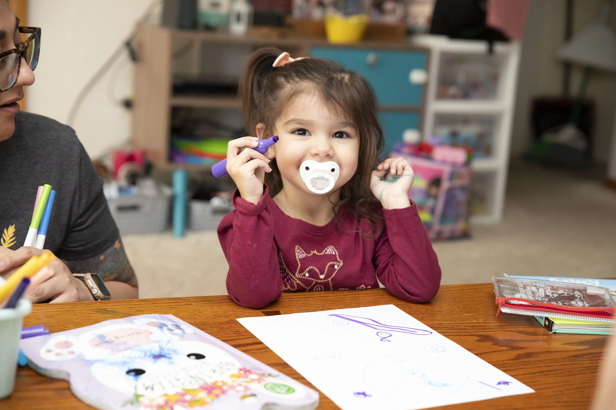A young girl with brown hair wearing a maroon long-sleeved shirt and a pink bow in her hair is sitting at a wooden table. She has a pacifier in her mouth, is holding a purple marker, and is smiling at the camera. There are colorful drawings on white paper in front of her, and various art supplies are scattered on the table. In the background, an adult wearing glasses and a gray shirt is partially visible.