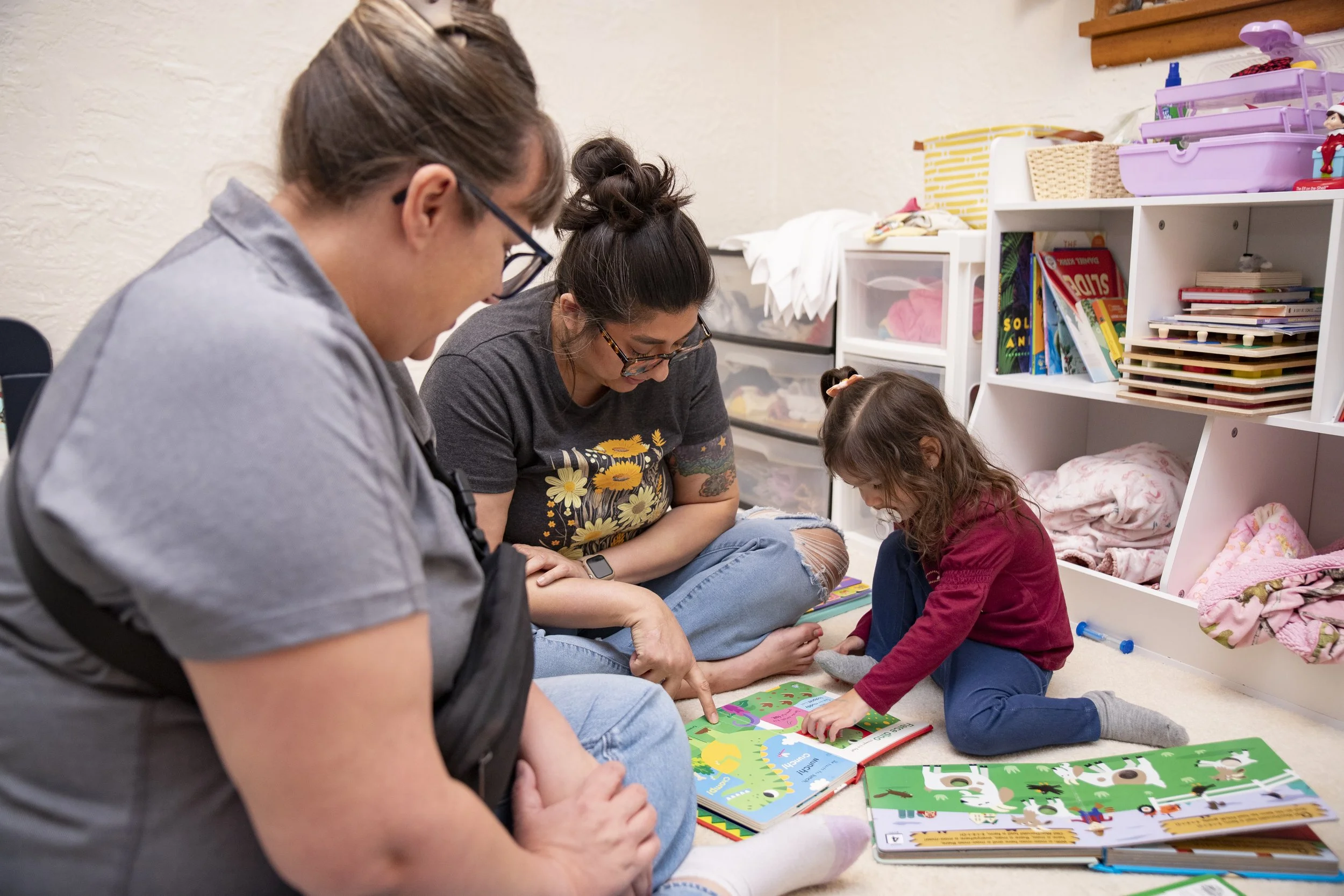 Three people, a woman, a man, and a young girl, sitting on the floor surrounded by books and reading to the girl, who is looking at a picture book.