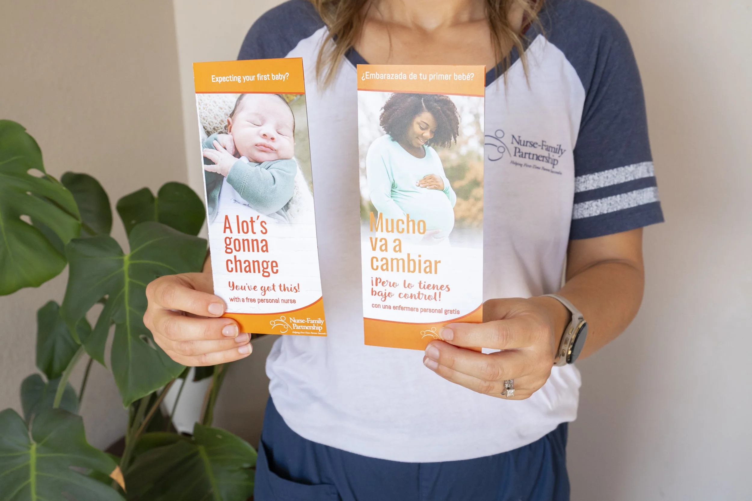 Woman holding two flyers from Nurse Family Partnership, one in English and one in Spanish, about pregnancy and newborn care, in front of a green plant.