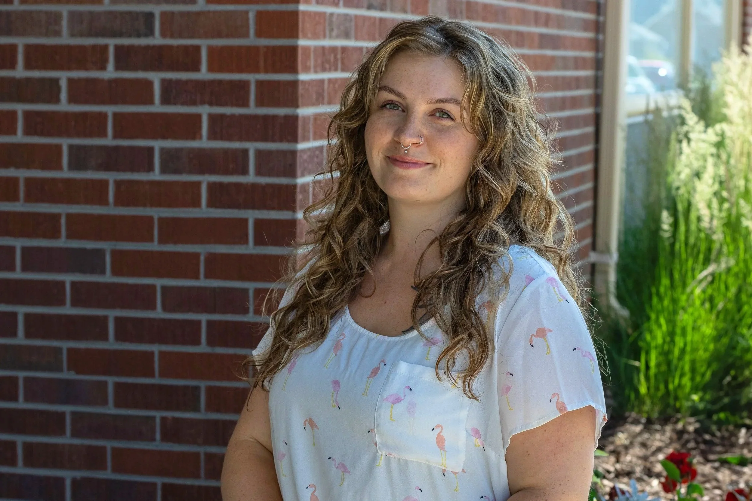 A woman standing outdoors near a brick wall, wearing a white shirt with flamingo prints, with her curly hair and a nose piercing, smiling slightly.