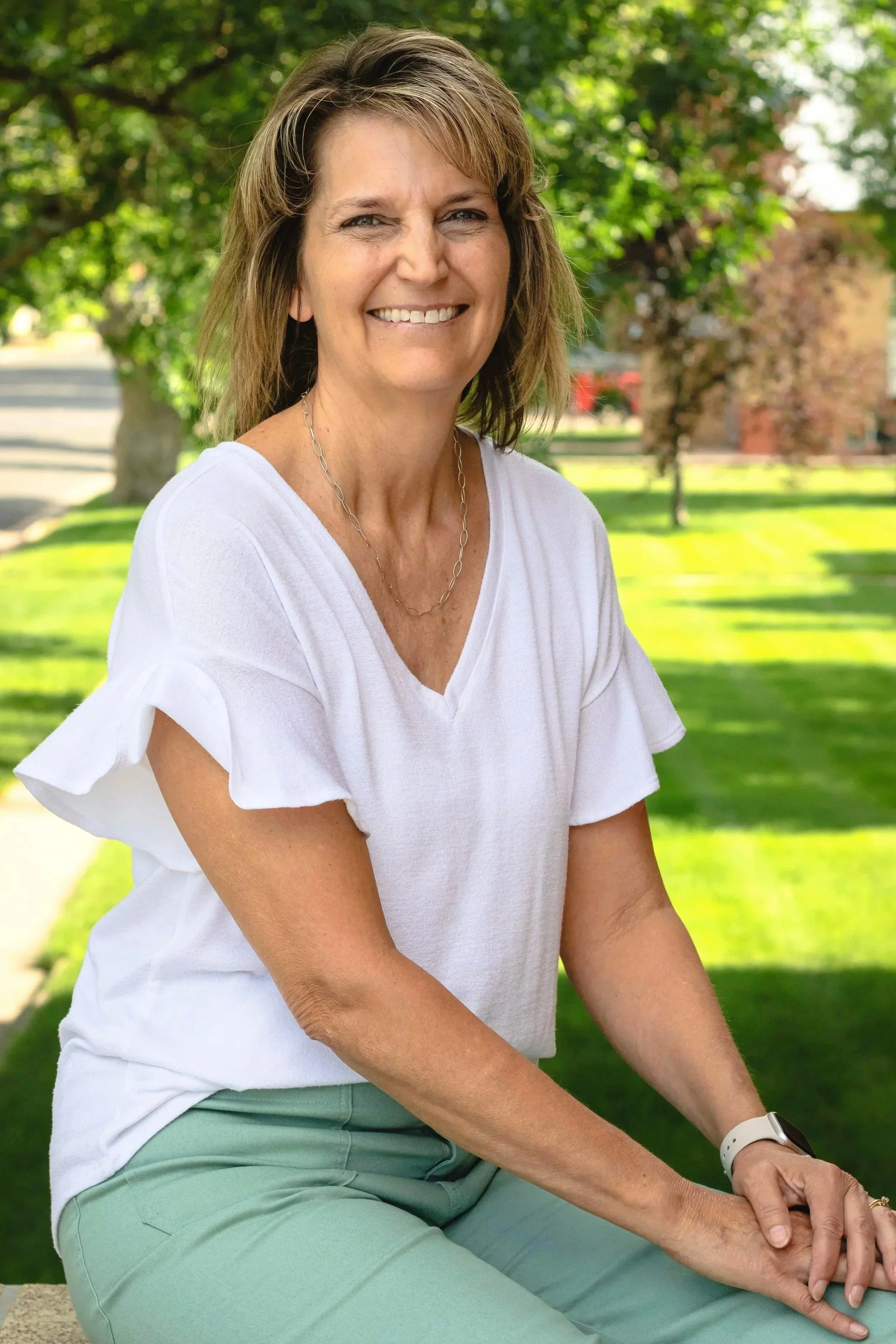 A woman smiling and sitting outdoors in a park with green grass and trees in the background, wearing a white top and light green pants.