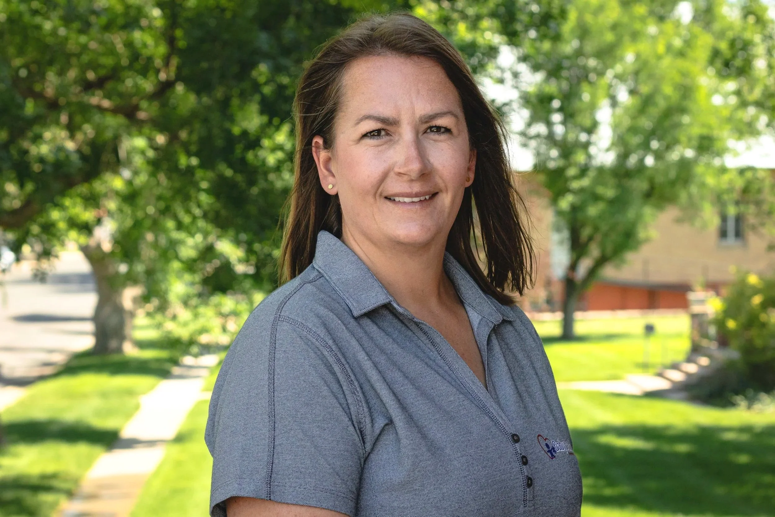 A woman with shoulder-length dark brown hair, wearing a gray collared shirt with a logo, standing outdoors on a sunny day with green trees and a residential neighborhood in the background.