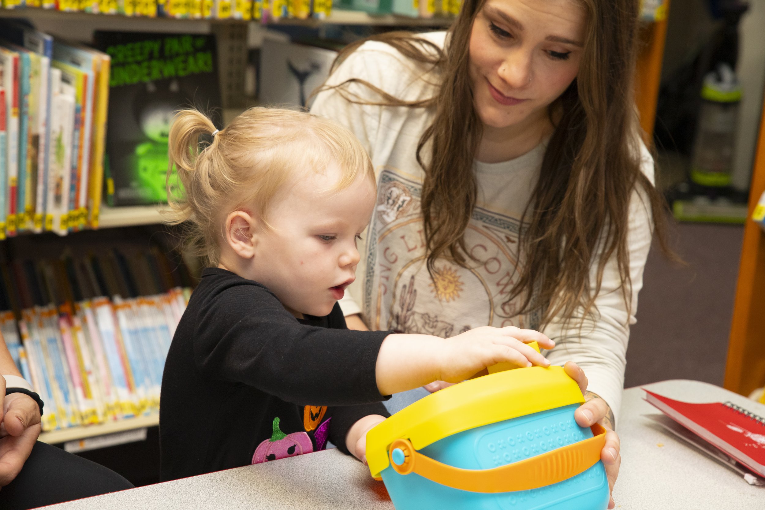 A young girl and woman are looking at a yellow and blue toy bucket together in a library or bookstore.