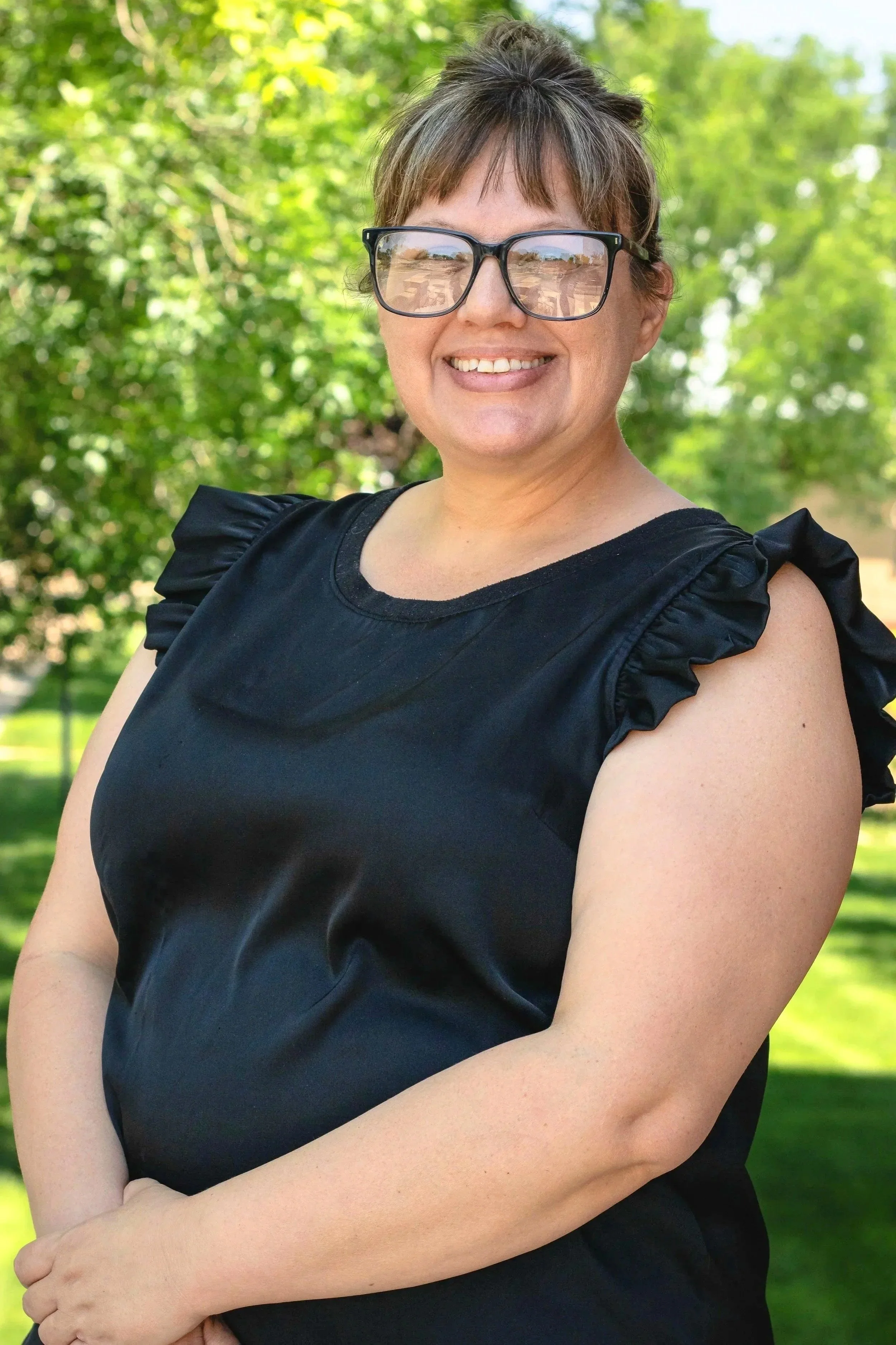 A smiling woman with short brown hair, wearing glasses and a black top with ruffled sleeves, standing outdoors with green trees in the background.