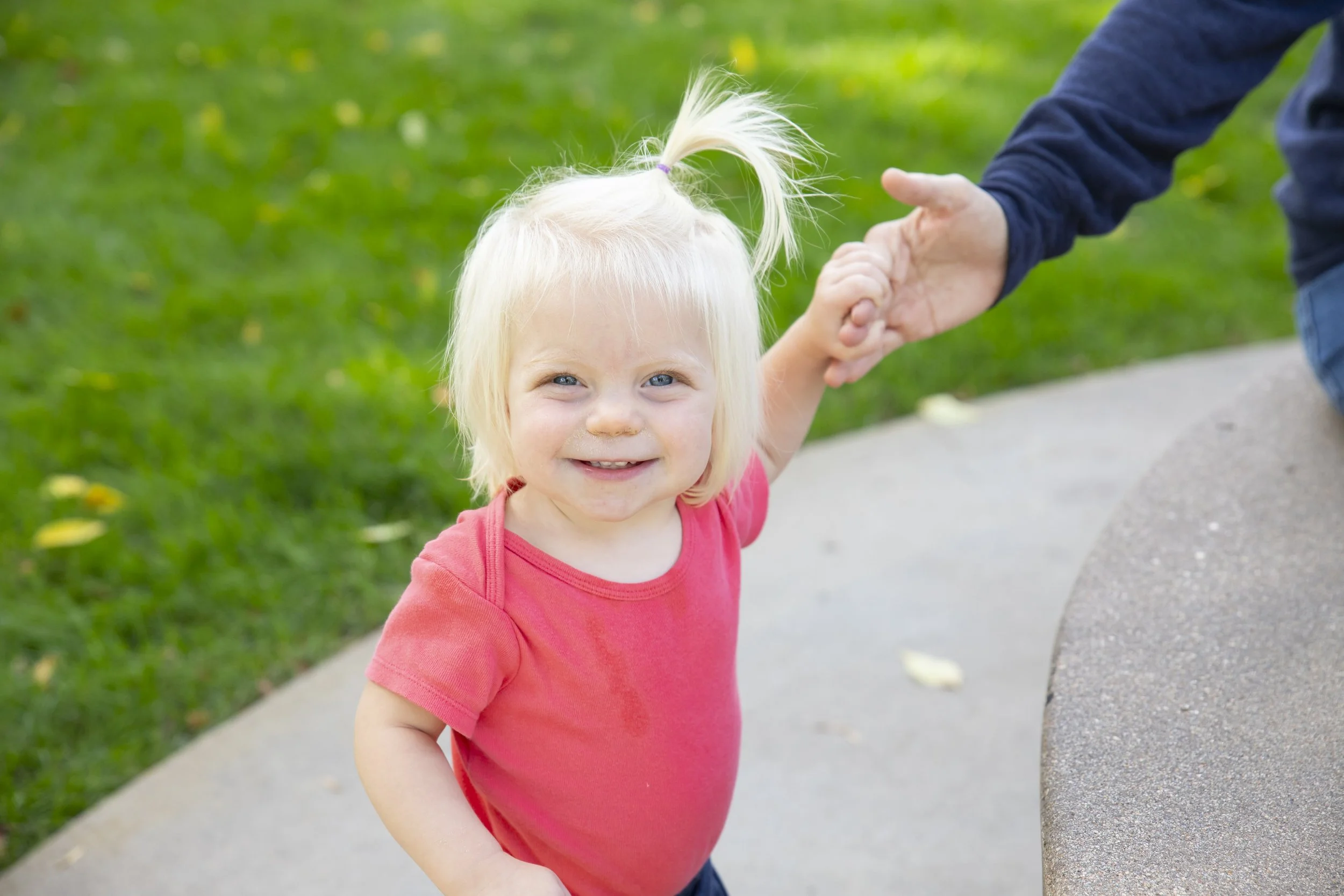 A young blonde girl in a pink shirt smiling while holding hands with an adult outside on a sidewalk with grass in the background.