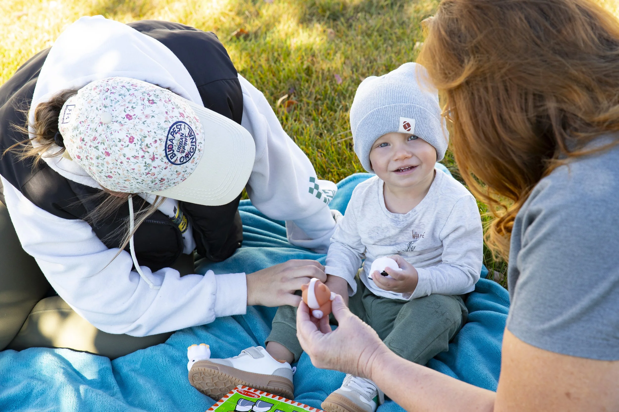 A young boy sitting on a blue blanket outdoors, smiling at the camera while holding a small object, with two women attending to him, one adjusting his shoe and the other holding a small item. The boy wears a gray beanie and a light-colored shirt. The women wear casual clothing and appear to be helping or playing with the boy.