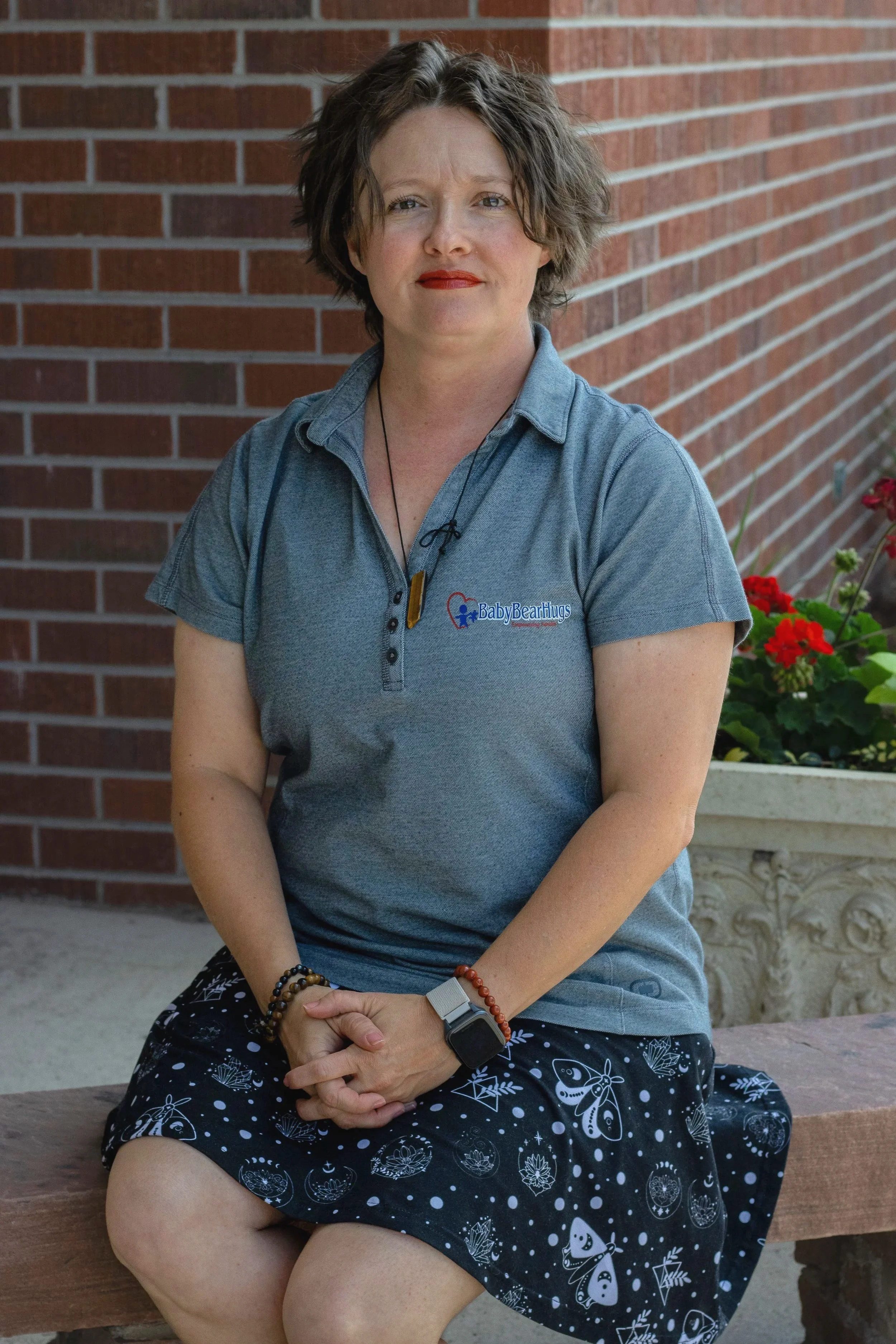 A woman sitting on a wooden bench wearing a gray polo shirt with a logo that reads "Baby Bear Hugs" and a black skirt with white butterfly and floral patterns, in front of a brick wall with red flowers.