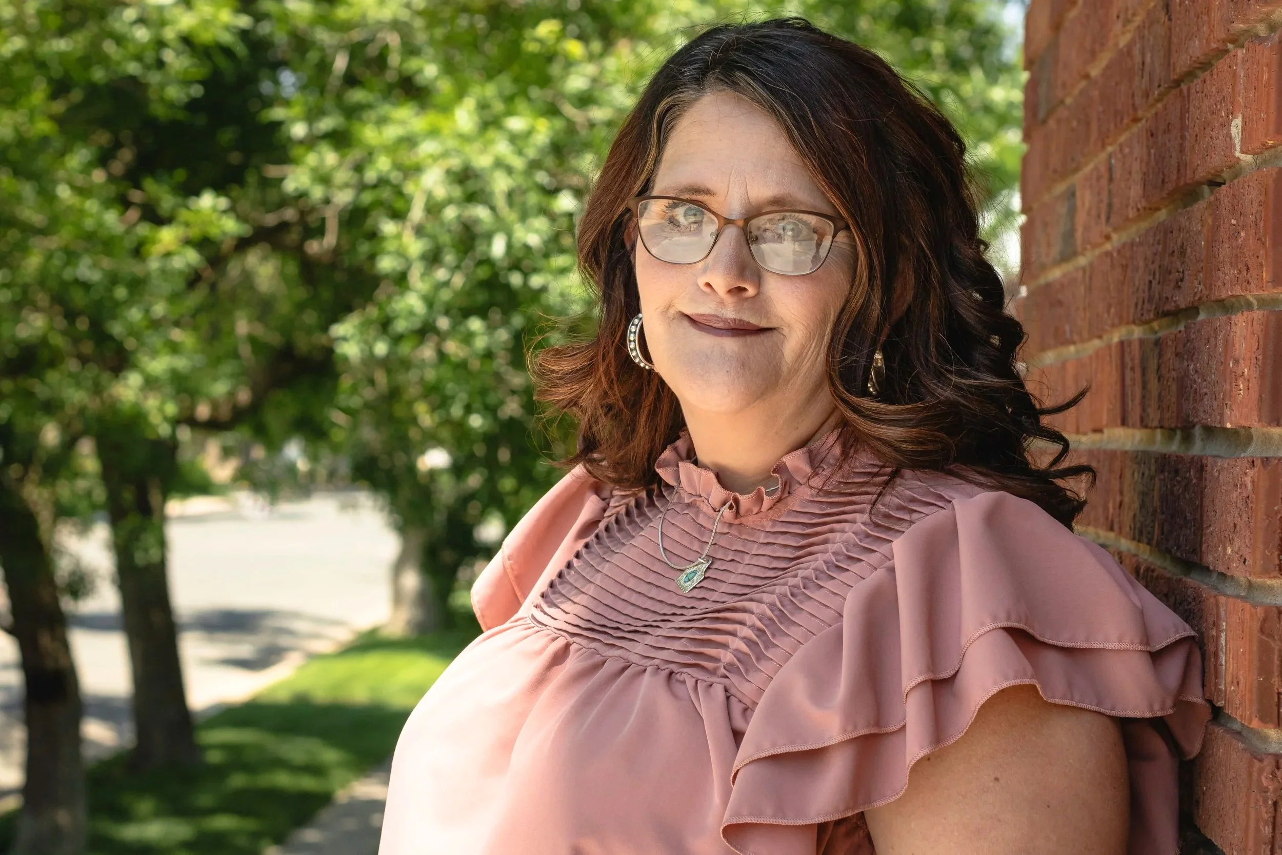 A woman with glasses and dark brown hair leaning against a brick wall outdoors, wearing a pink ruffled top and jewelry, with trees and a sidewalk in the background.