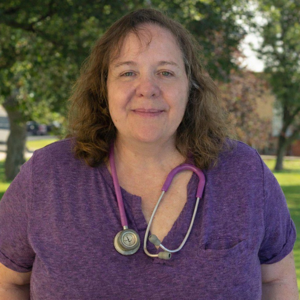 A middle-aged woman with long, wavy gray hair and glasses, wearing a black shirt and jacket, outdoors in a green park with trees and a pathway in the background.