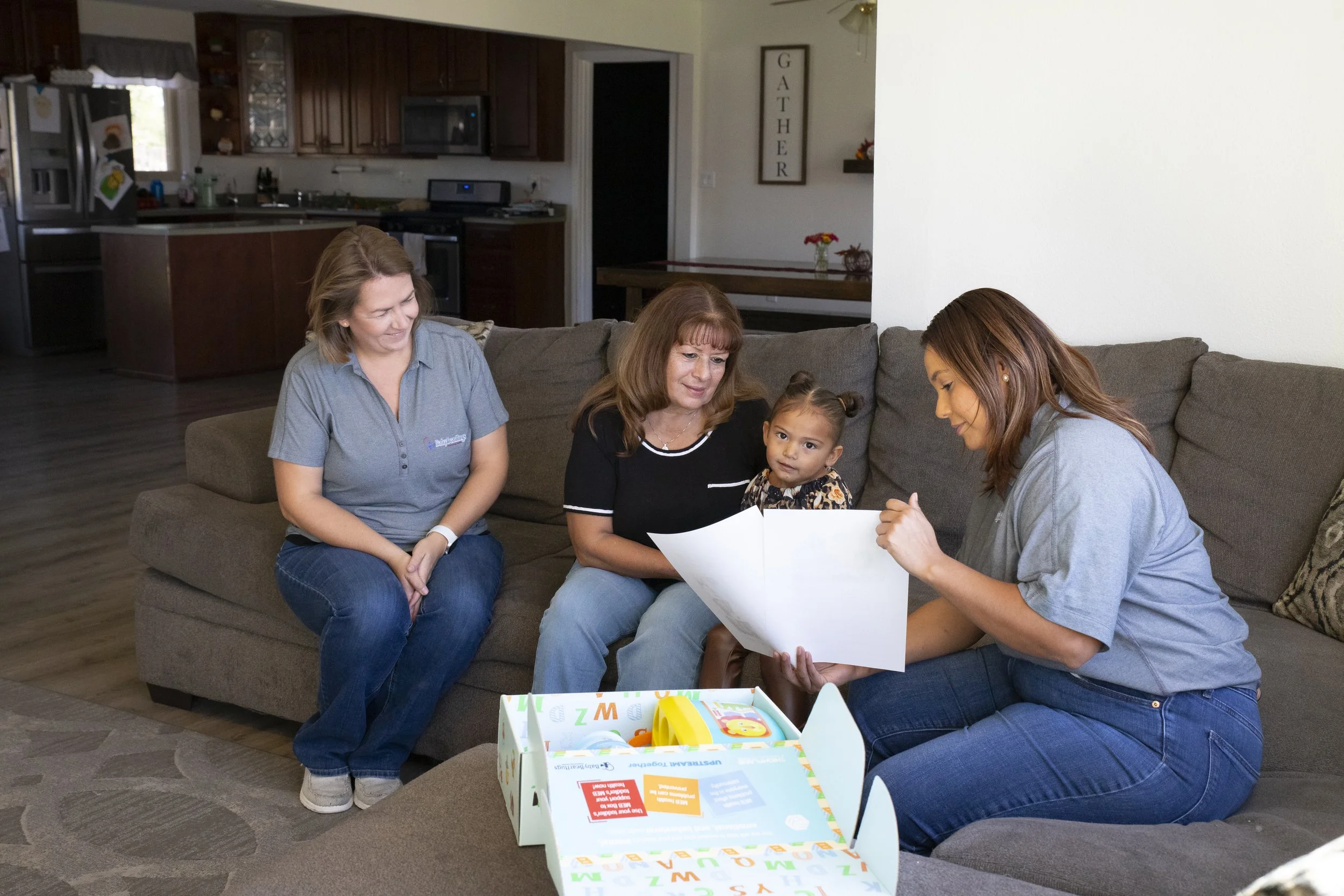 A group of four women and one young girl sitting together on a brown couch in a living room, looking at a large piece of paper or a book, with an open colorful box on the table in front of them.