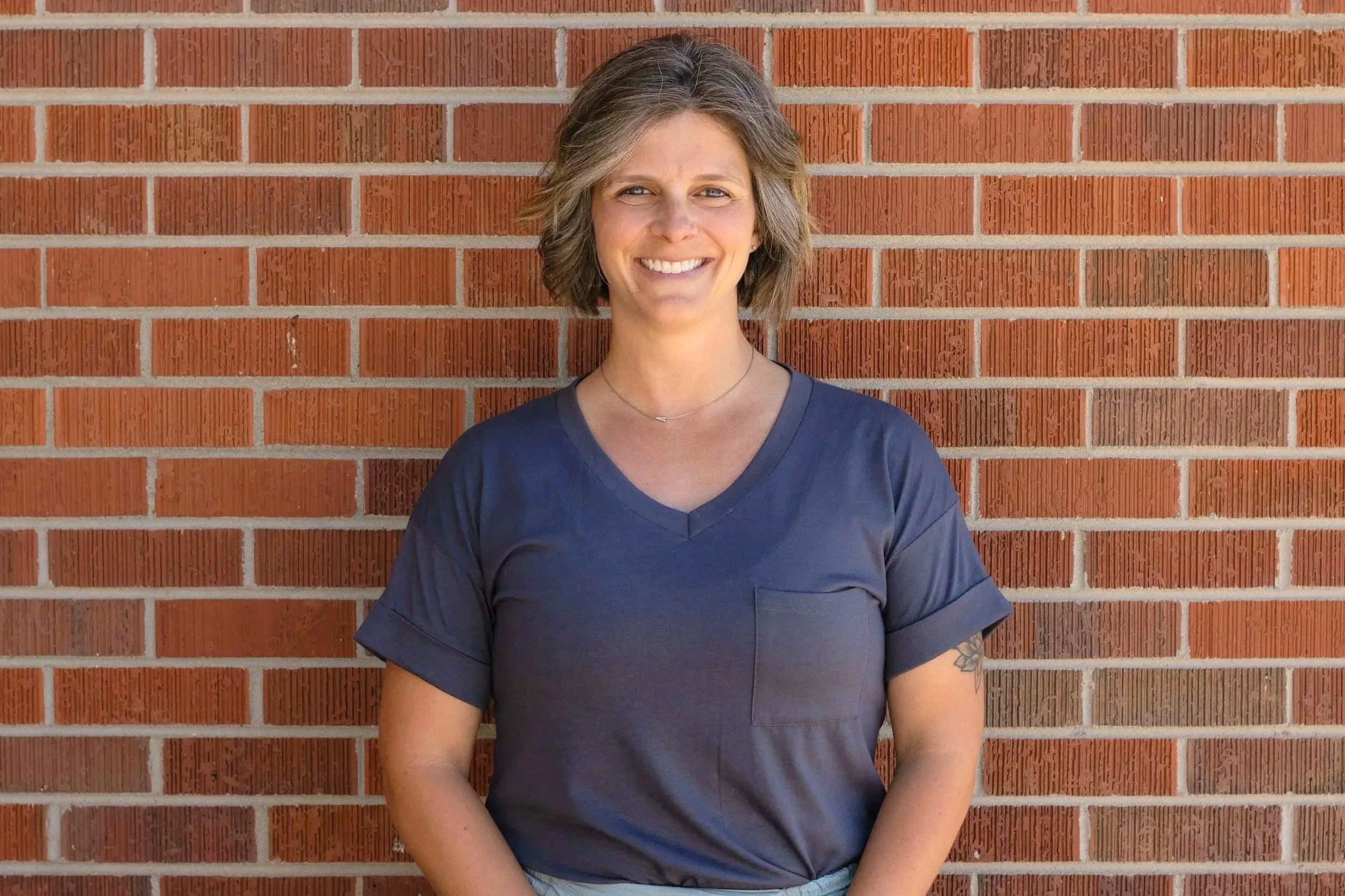 A woman with short brown hair smiling, standing against a red brick wall, wearing a dark blue V-neck T-shirt.