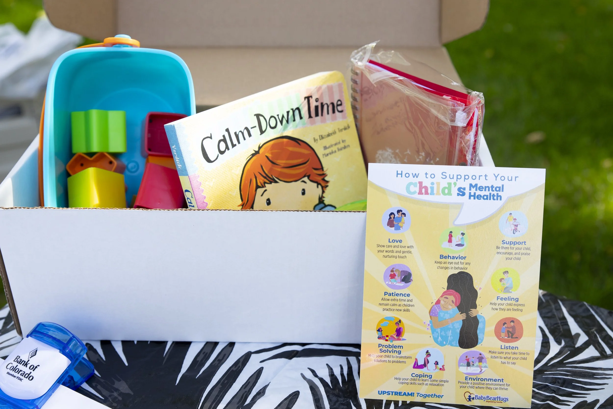 A box containing children's items including a book titled 'Calm-Down Time,' colorful toy building blocks, and a plastic container. A brochure titled 'How to Support Your Child's Mental Health' with tips for love, support, behavior, and more, stands outside the box. A small blue tape dispenser labeled 'Bank of Colorado' is also visible.