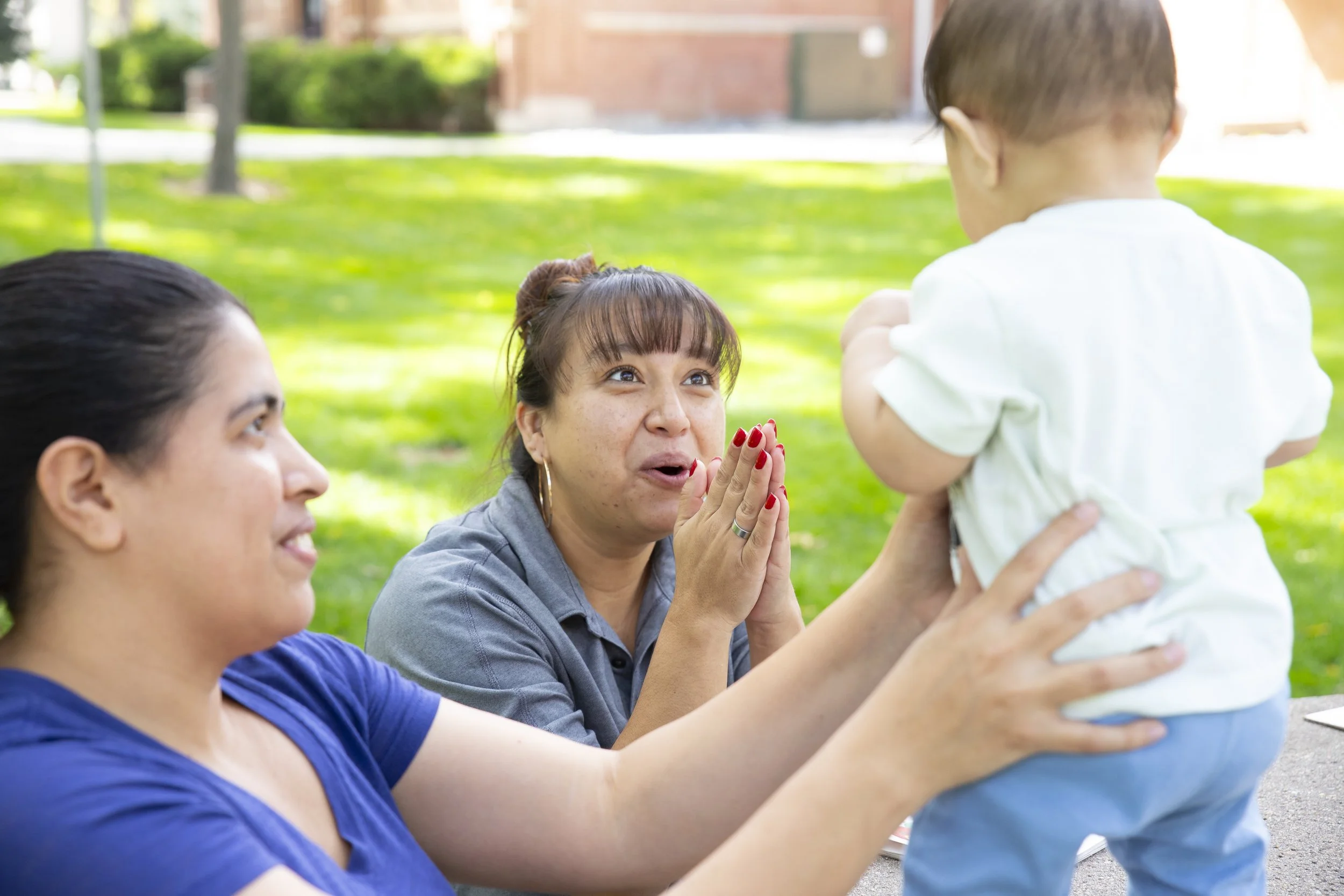 Two women and a young child outdoors on a sunny day, with the woman in gray excitedly clapping and the woman in blue smiling as they interact with the child.