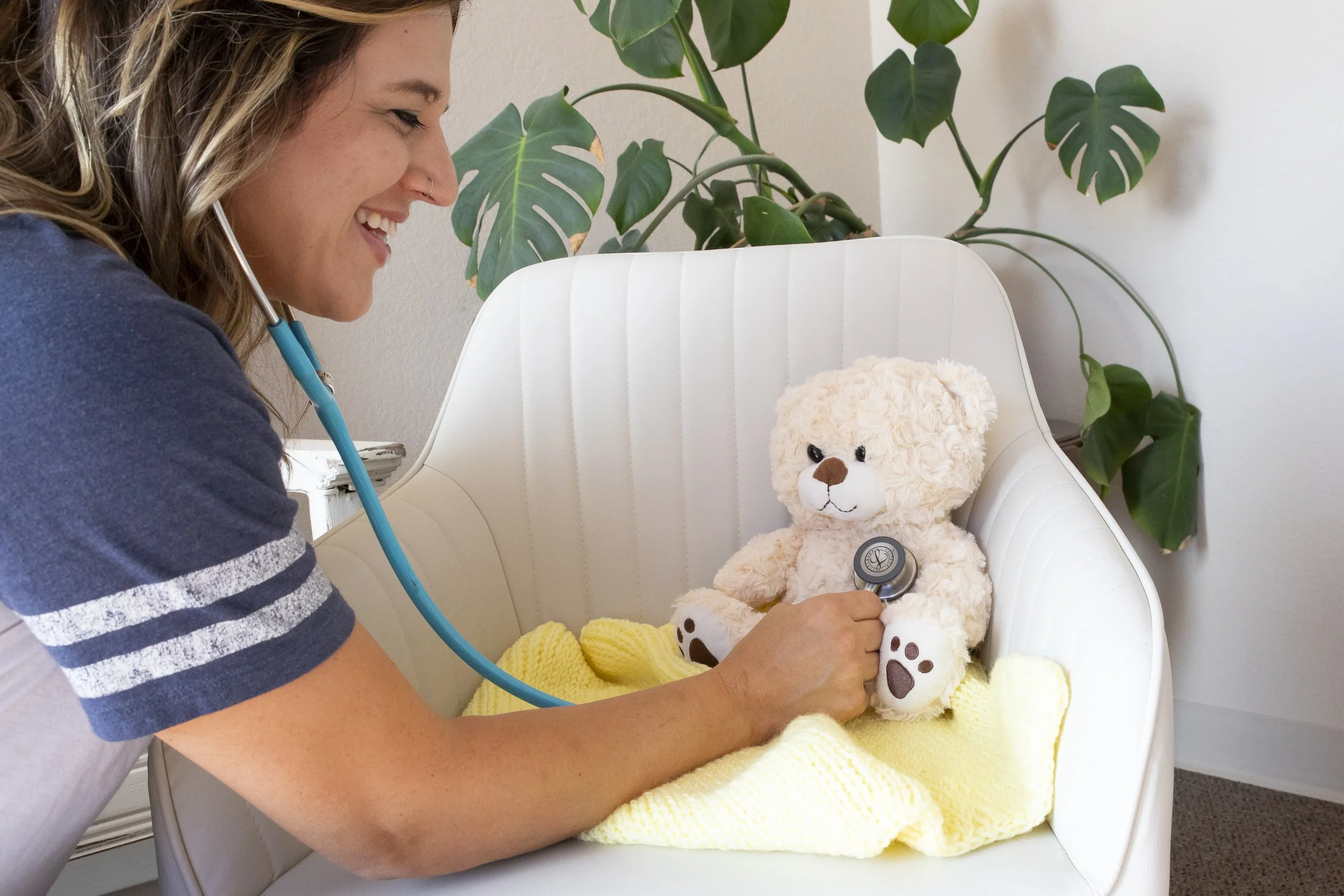 A woman with a stethoscope listening to a teddy bear that is seated on a white chair with a yellow blanket, in front of a green leafy plant.