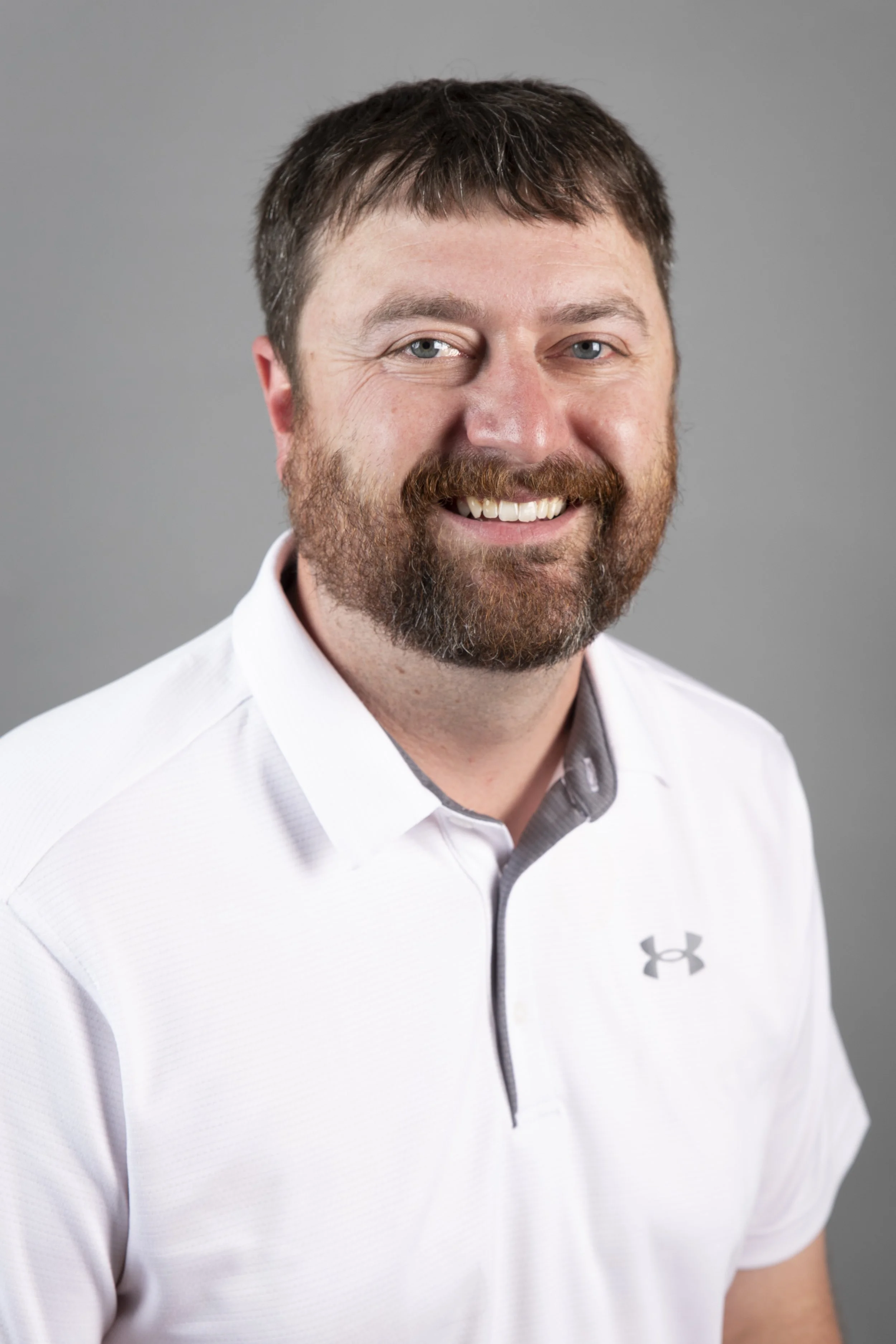 Portrait of a smiling man with brown hair and beard, wearing a white collared shirt with a gray zipper and Under Armour logo on the chest, against a gray background.