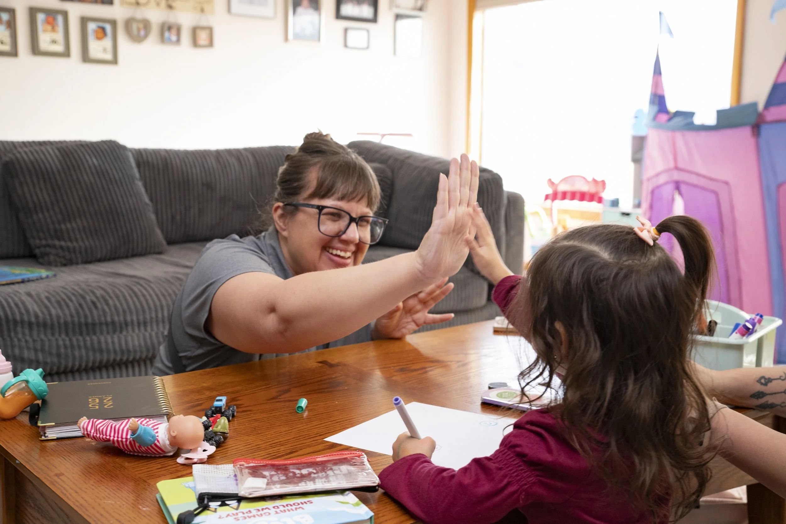 A woman and a young girl sharing a high-five at a wooden table. The woman has glasses and brown hair, smiling. The girl has curly hair with a hair clip, holding a marker, with coloring supplies and a doll on the table.