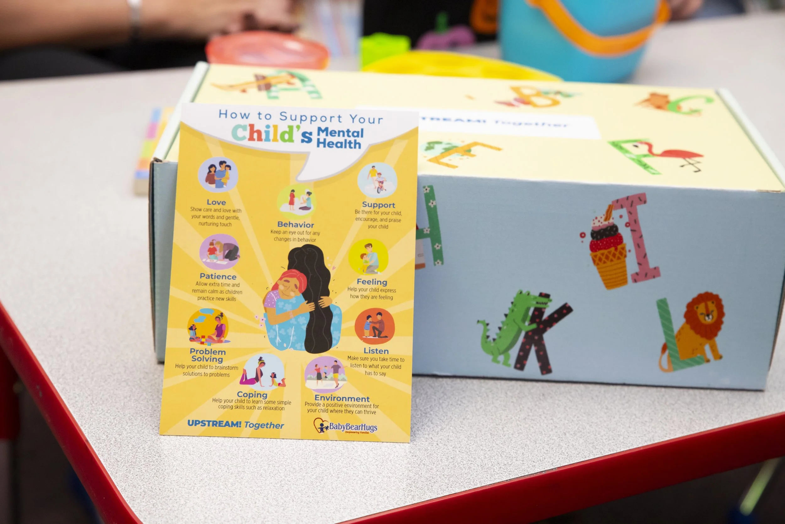 A colorful pamphlet titled 'How to Support Your Child's Mental Health' with tips such as love, patience, problem solving, coping, support, and environment. The pamphlet features an illustration of a child hugging an adult. The pamphlet is in front of a blue box decorated with letter and animal cutouts, on a school table.