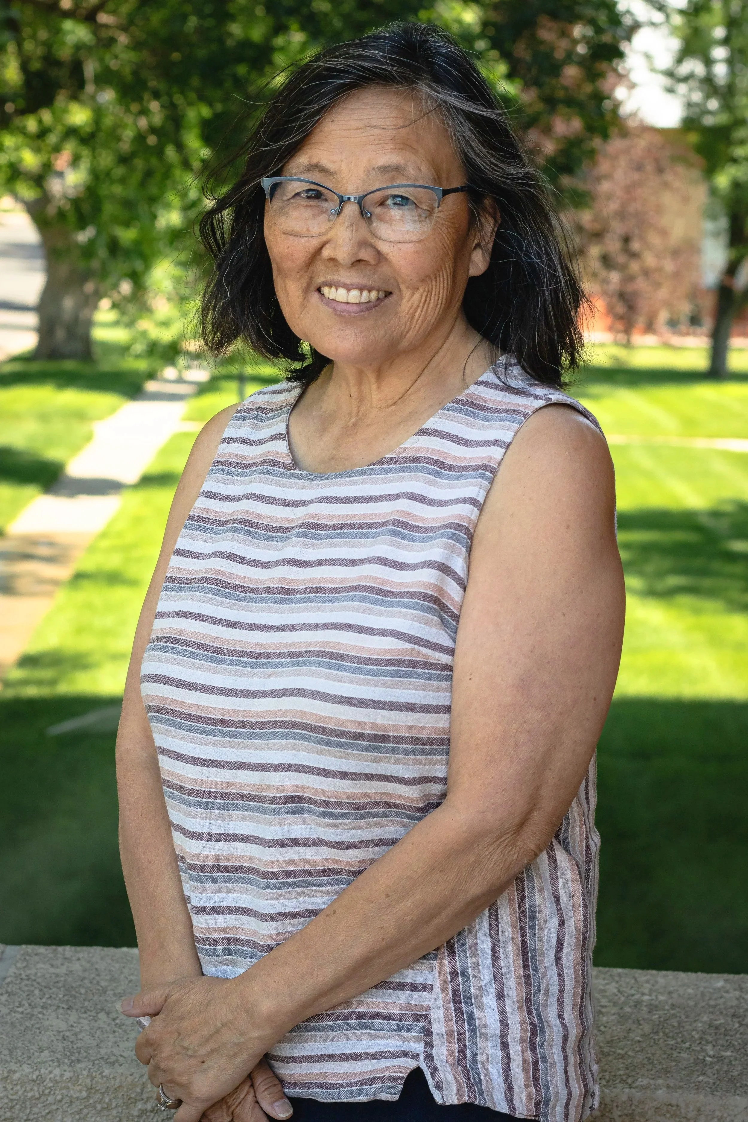 An elderly woman with glasses and shoulder-length black hair standing outdoors in a park during daytime, smiling, wearing a sleeveless striped top.