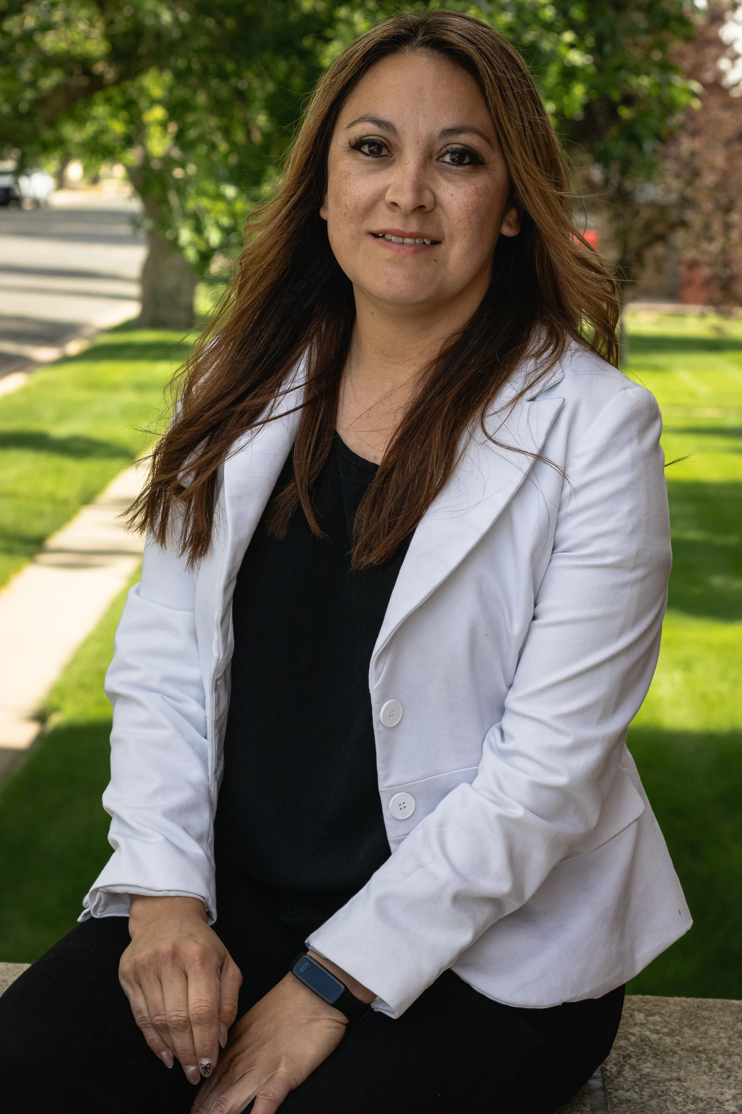 A woman with long brown hair, wearing a white blazer and black shirt, sitting outdoors on a stone surface with a grassy lawn and trees in the background.