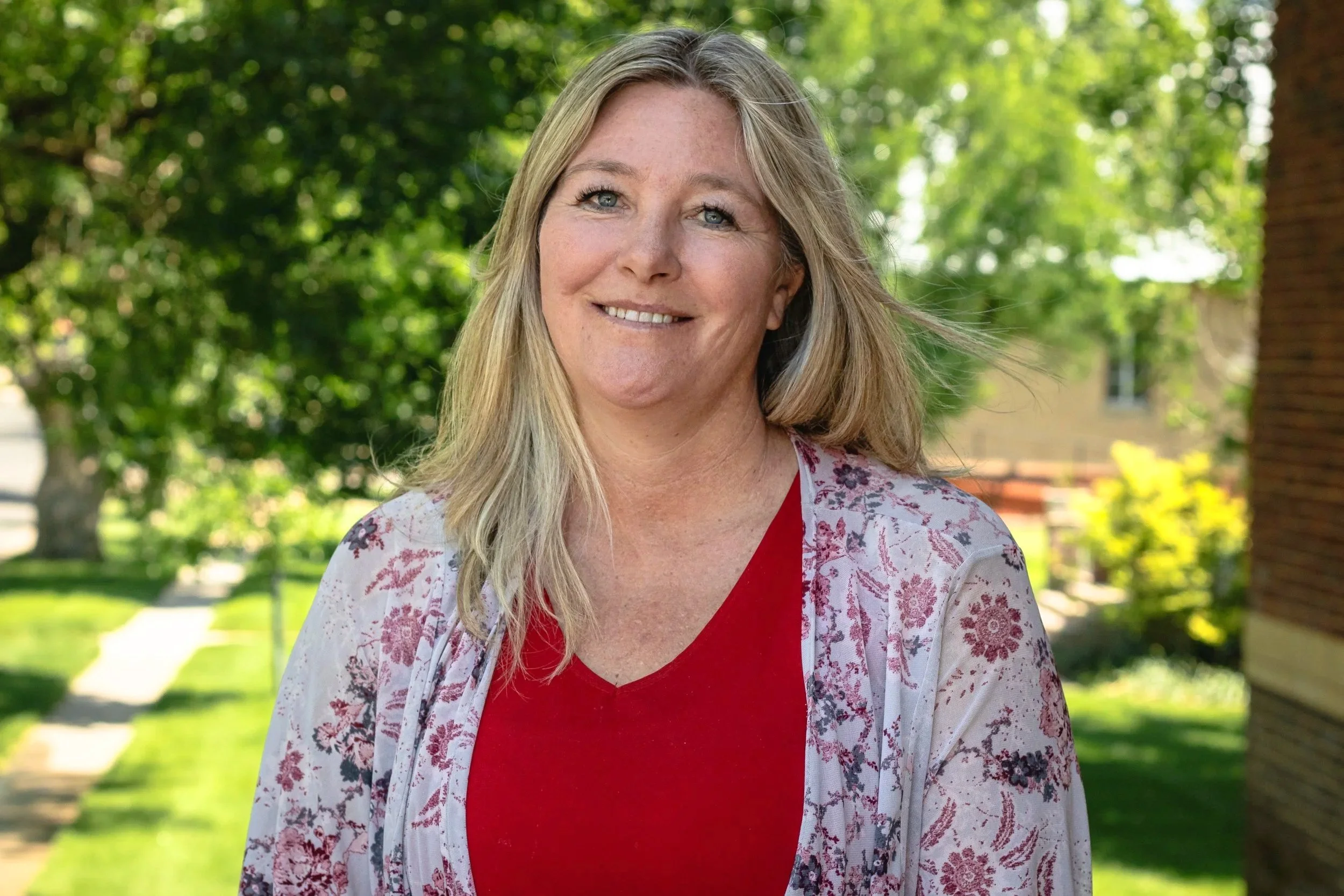 A smiling woman with blonde hair standing outside on a sunny day with green trees and a sidewalk in the background.