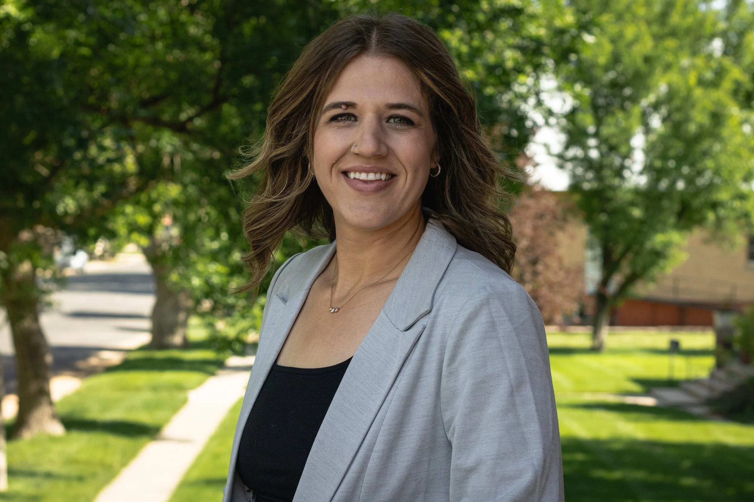 A woman with wavy brown hair smiling outdoors on a sunny day with trees and a sidewalk in the background.