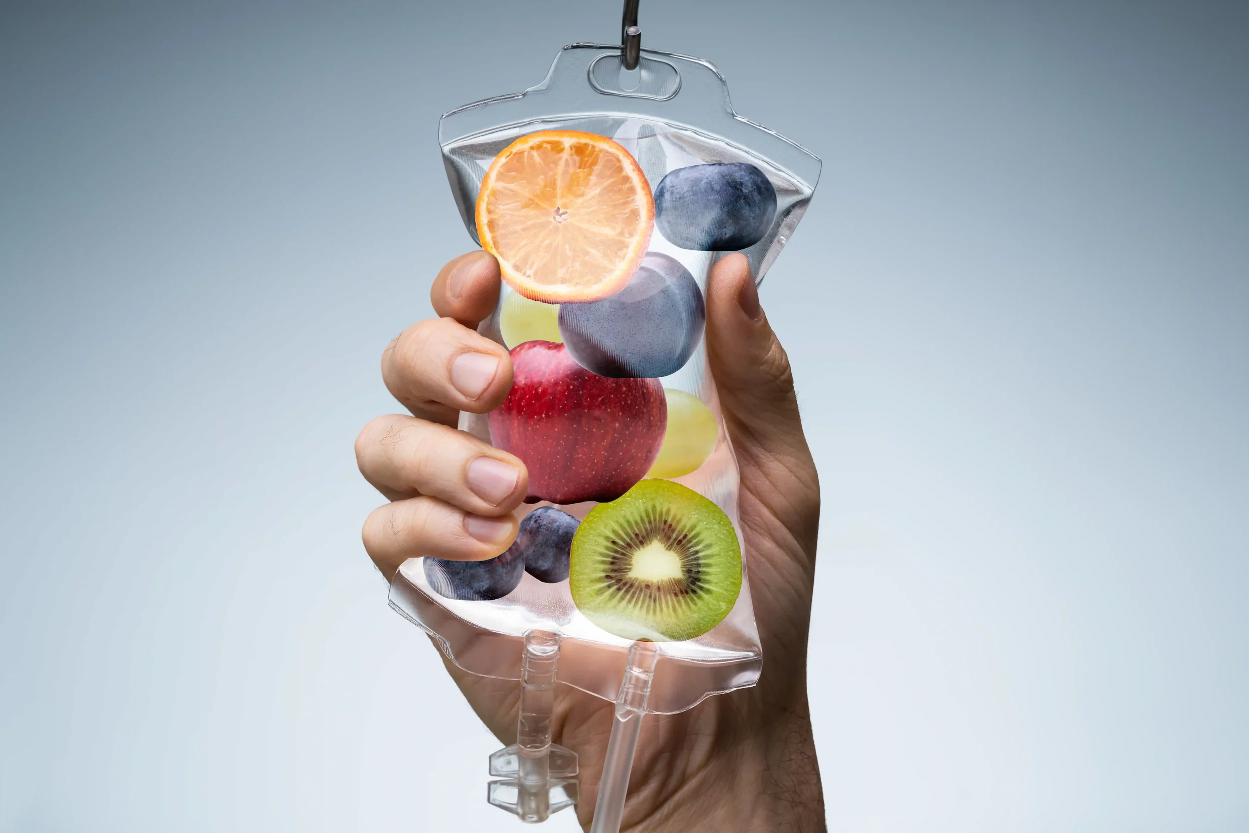 Hand holding a transparent IV bag with images of various fruits like orange, blueberry, apple, kiwi, and grapes inside.