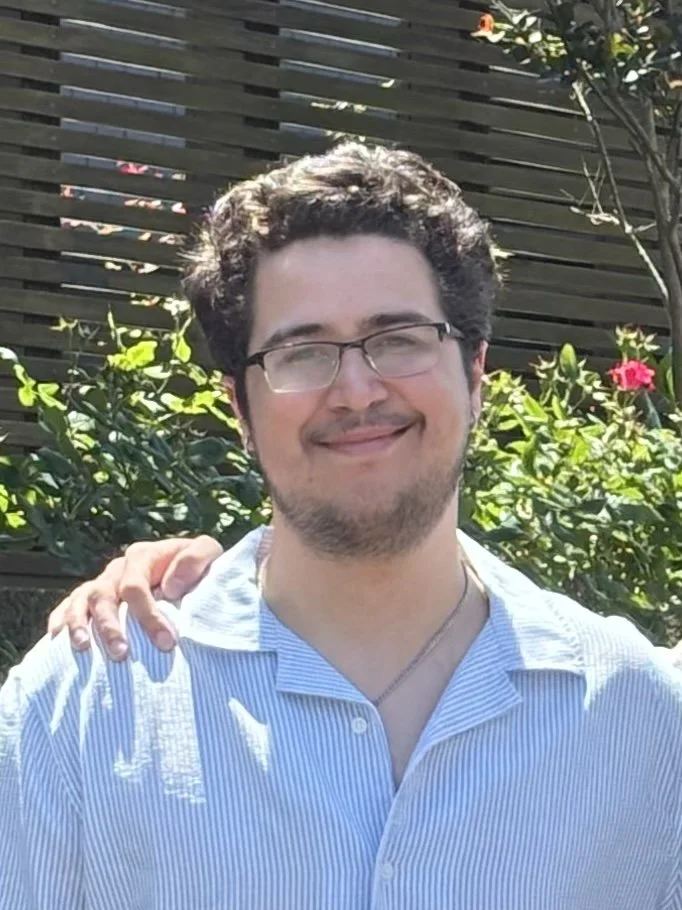 A young man with glasses and a beard smiling outdoors, with greenery and a wooden fence in the background.
