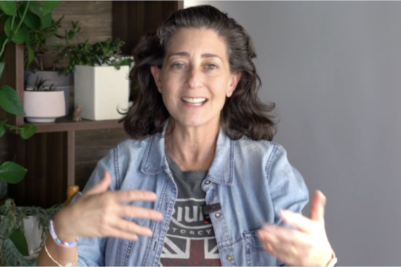 Woman with dark curly hair gesturing with hands and smiling, wearing a denim jacket and a t-shirt, in a room with plants and wooden shelves.