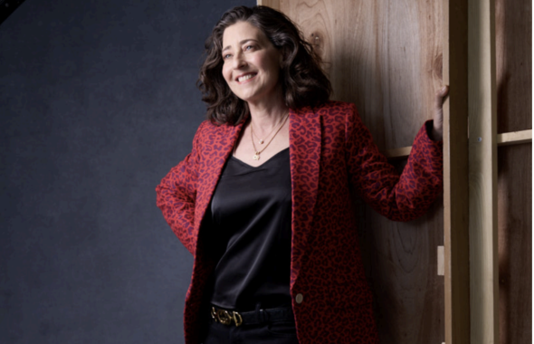 A woman with shoulder-length dark curly hair, smiling, wearing a red patterned blazer over a black blouse, standing against a wooden door and a dark wall.