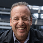 A smiling middle-aged man with short dark hair, wearing a dark jacket, standing outdoors with a stadium in the background.