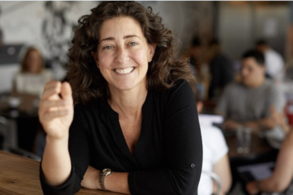 A woman smiling at a cafe or restaurant, with other people in the background.