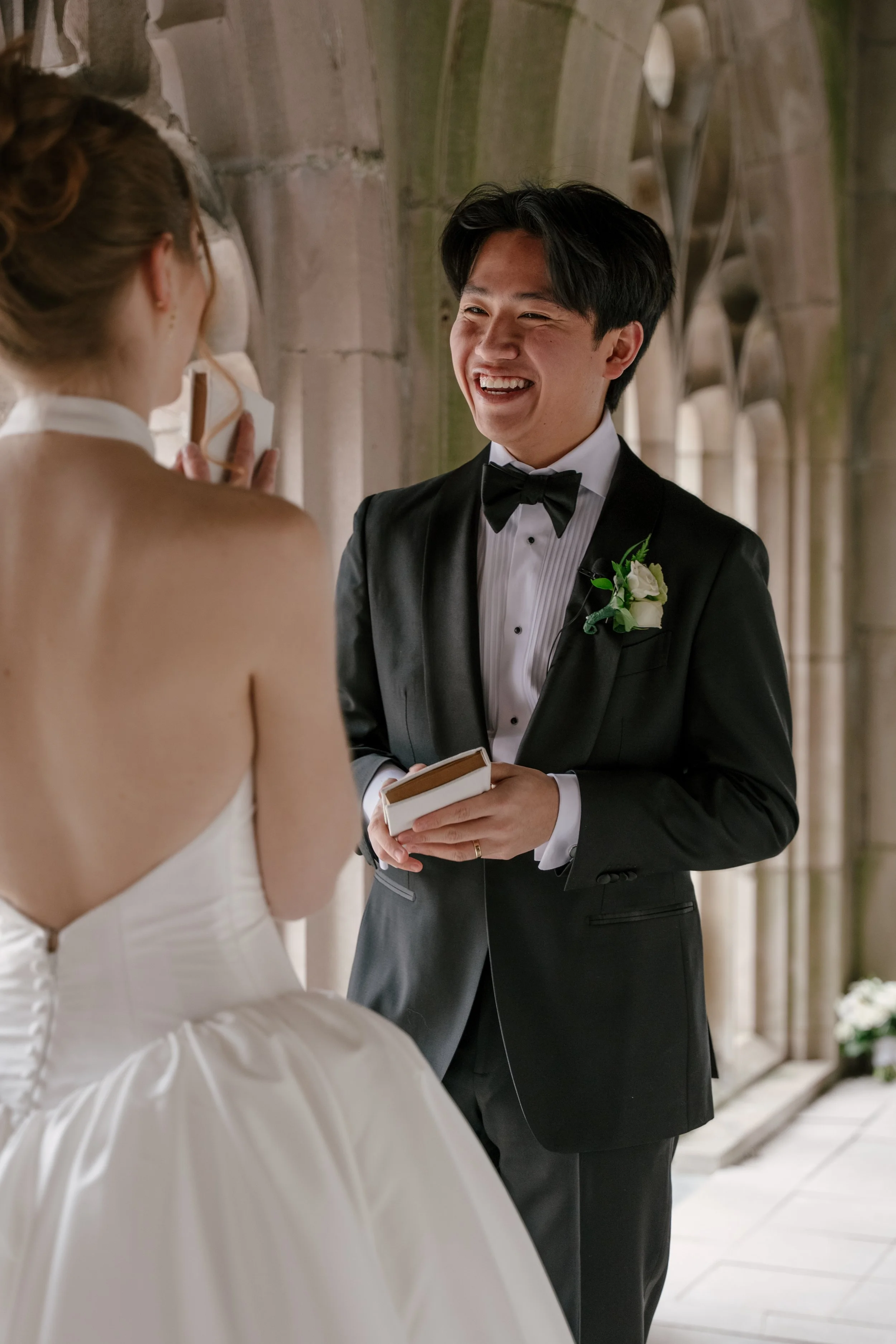 A groom in a black tuxedo with a boutonniere on his lapel is smiling and holding a small book in a church with stone arches. A bride in a strapless wedding gown with her hair styled up is seen from behind, holding a small object close to her ear, fac